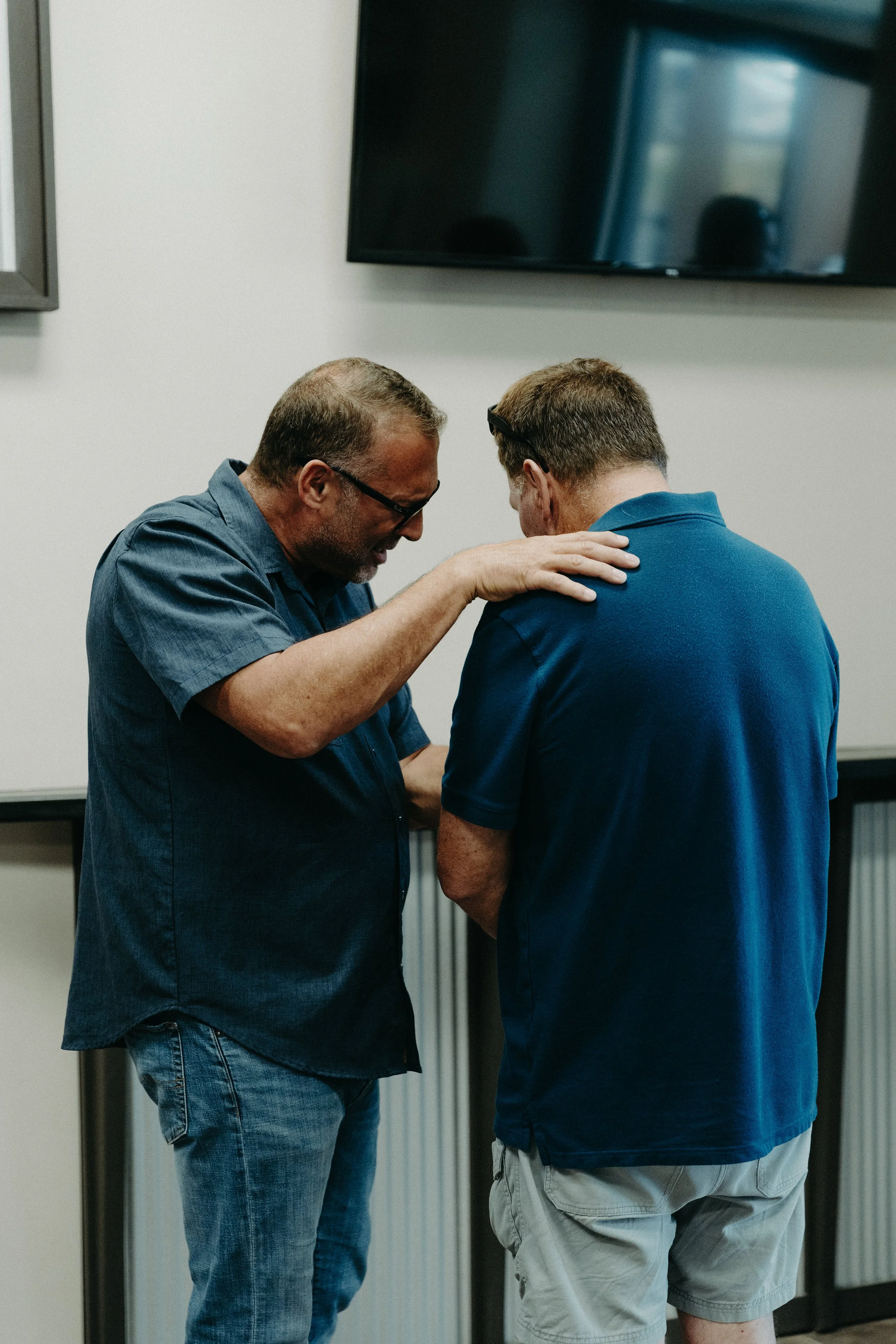 Two men are kneeling and praying together inside a room with a television mounted on a plain wall.