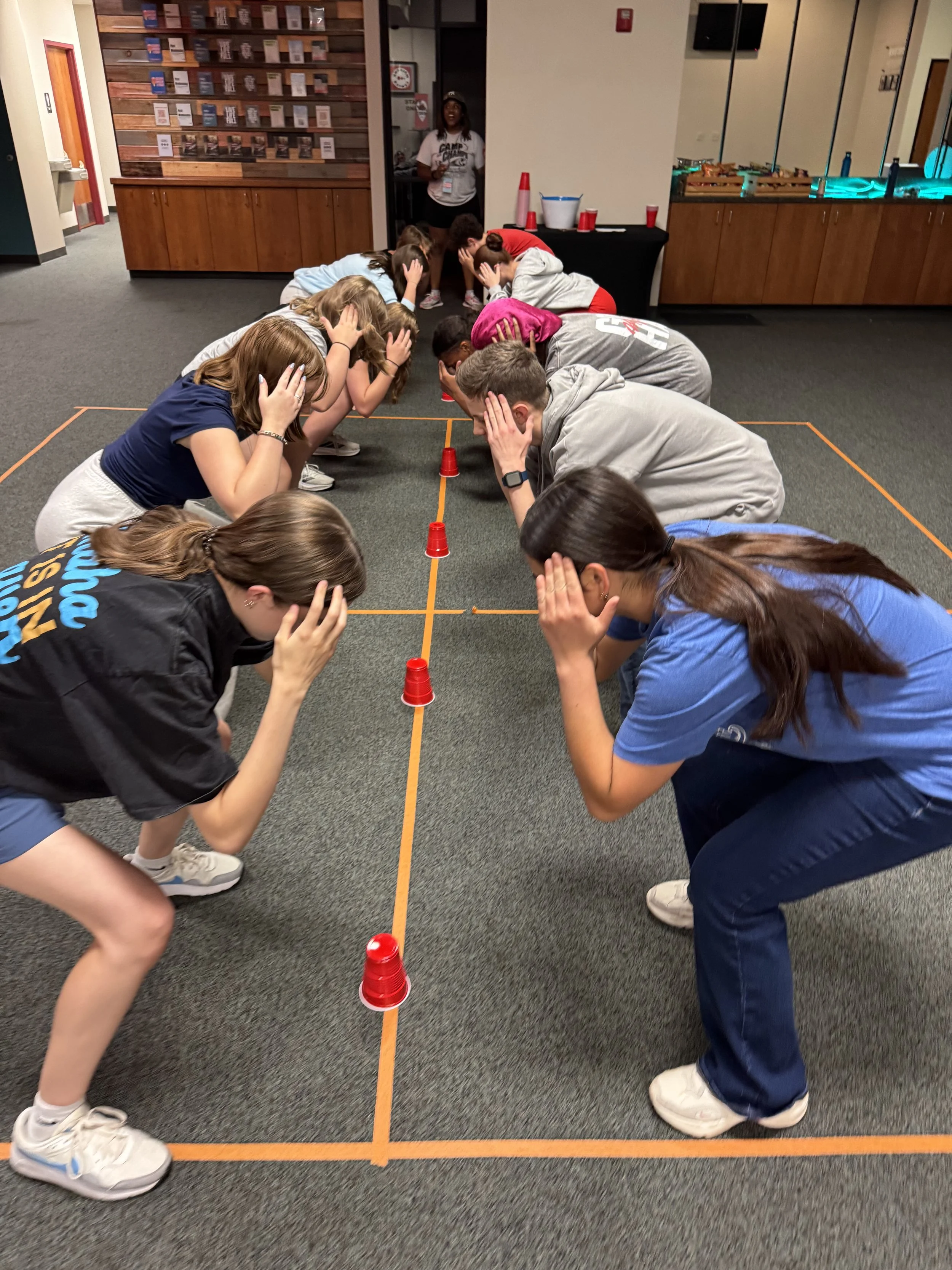 Group of children and teenagers participating in a game or activity that involves covering their ears while crouching in a line marked by orange tape, with red cups placed along the tape on a carpeted floor.