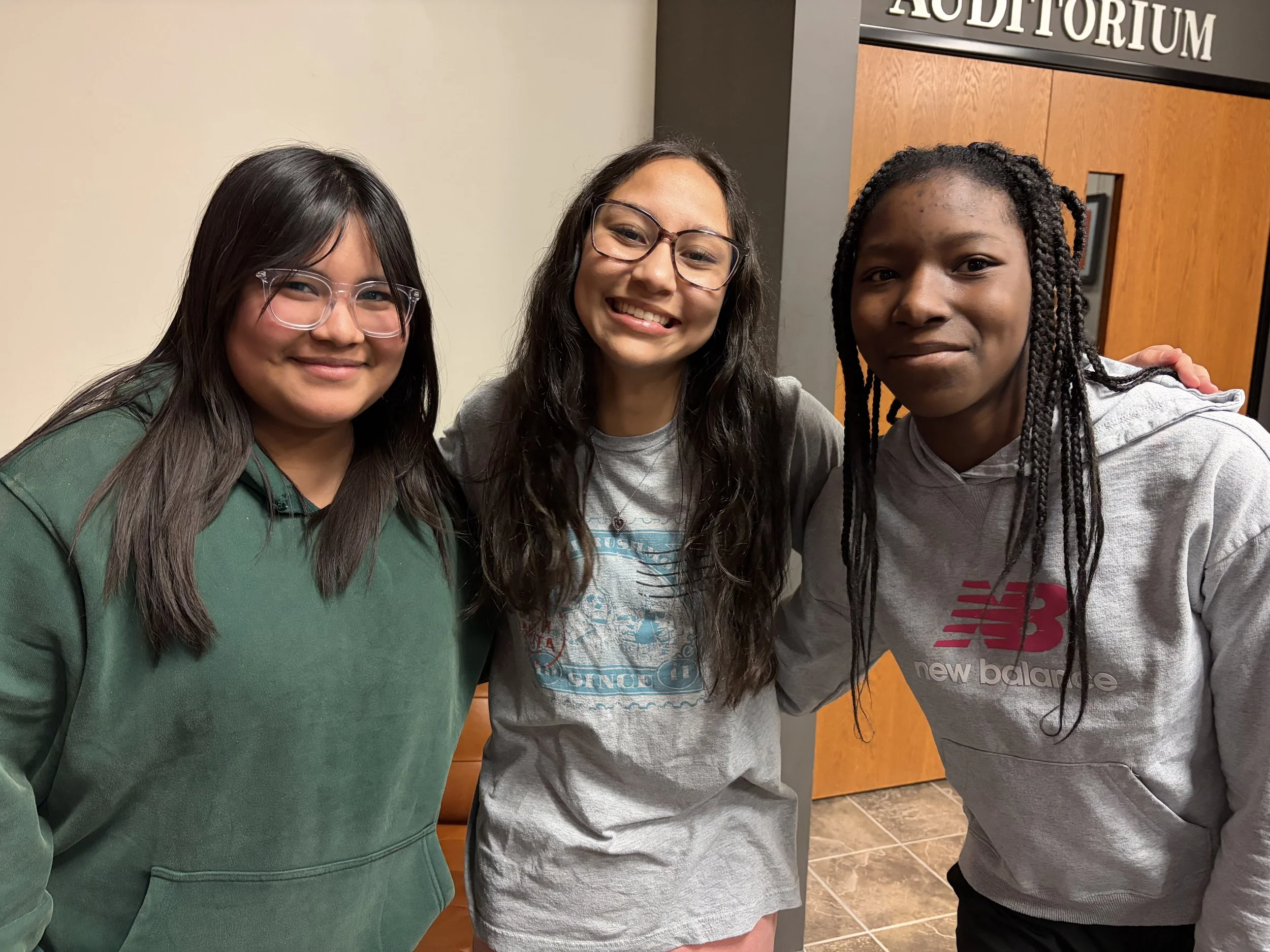 Group of three young girls standing close together, smiling, in an indoor location with a sign reading 'AUDITORIUM' in the background.