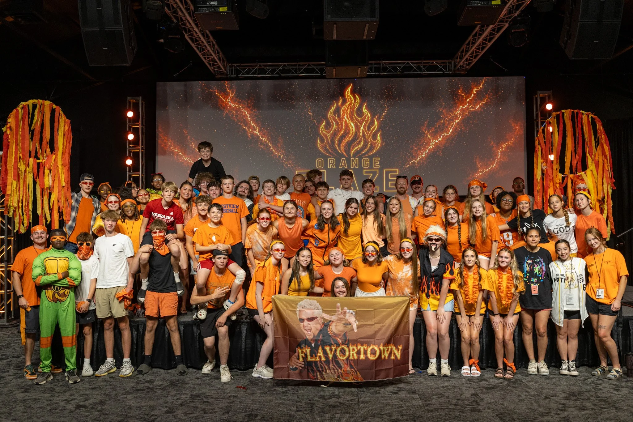 Group of children and adults gathered on stage at an event called 'Flavor Town,' with a large screen behind them displaying fiery graphics and the event name.