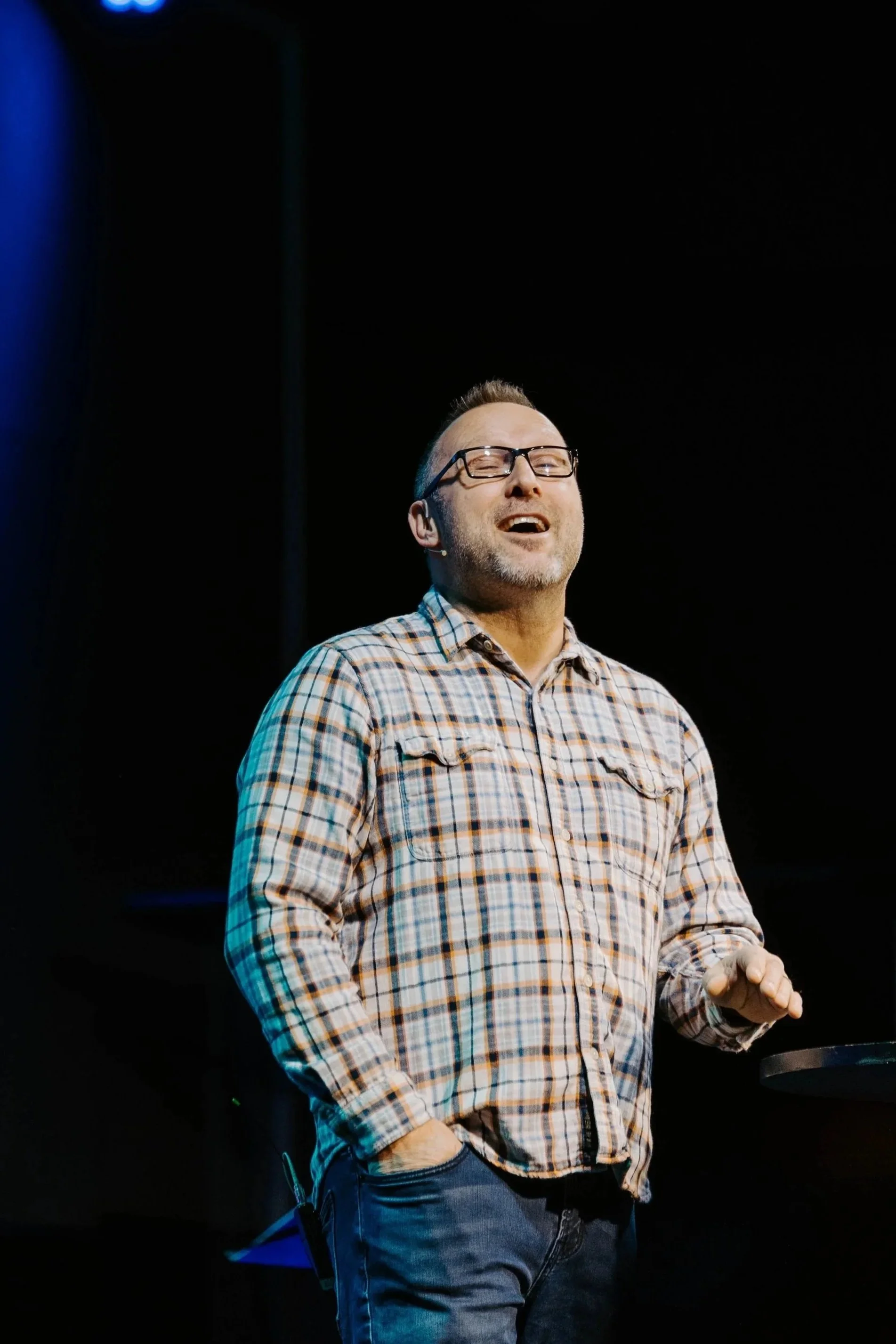 A man wearing glasses and a plaid shirt smiling and speaking on stage, with dark background and stage lighting.
