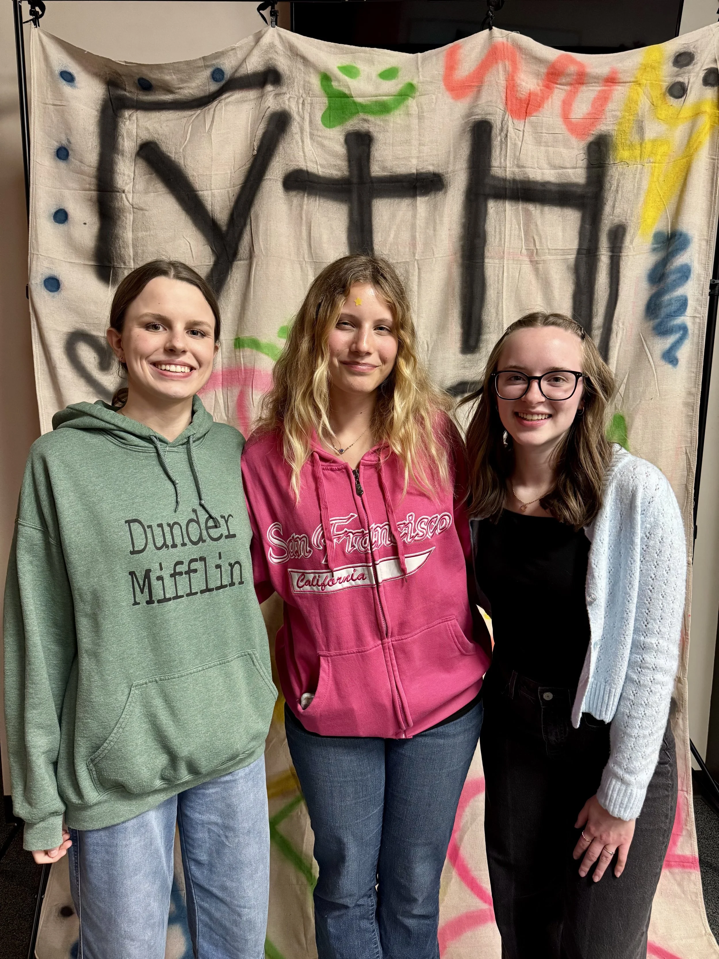 Three young women smiling, standing in front of a painted cloth backdrop with colorful graffiti, including words like 'Y' and 'Las'. The woman on the left is wearing a green hoodie with 'Dunder Mifflin' printed on it, the woman in the middle is weari