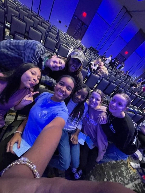 Group of six women posing for a selfie in an empty auditorium or theater with blue lighting.