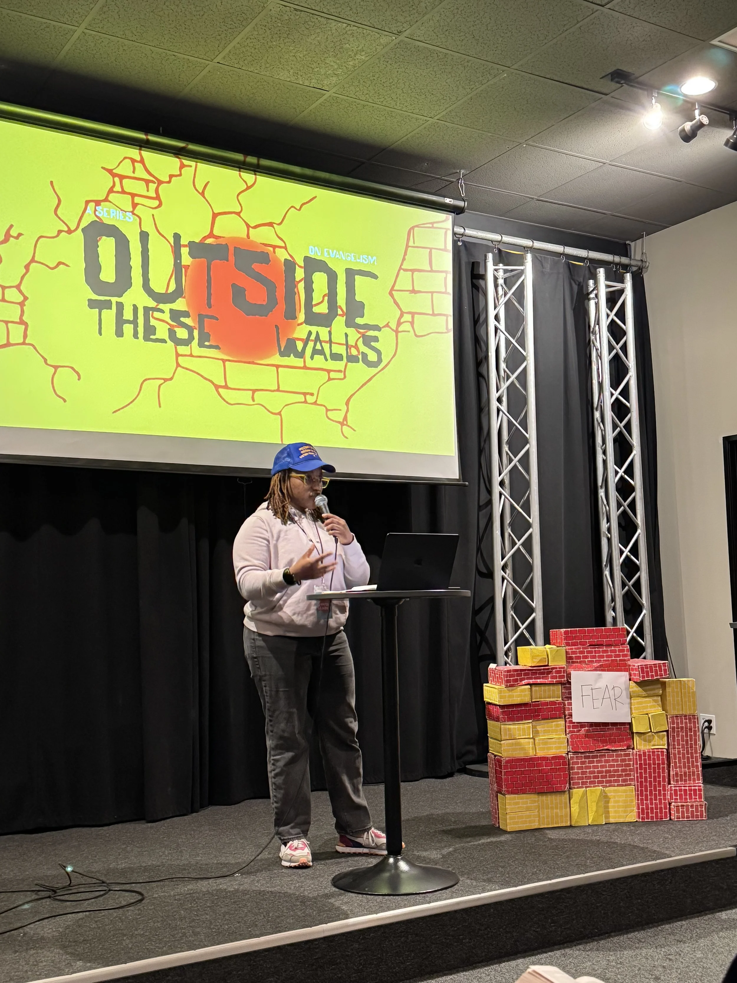 A woman speaking at a podium during a presentation or talk, with a large screen behind displaying text and graphics about 'Inside Walls' and evangelism. There are stacks of blocks labeled 'FEAR' to her right on the stage.