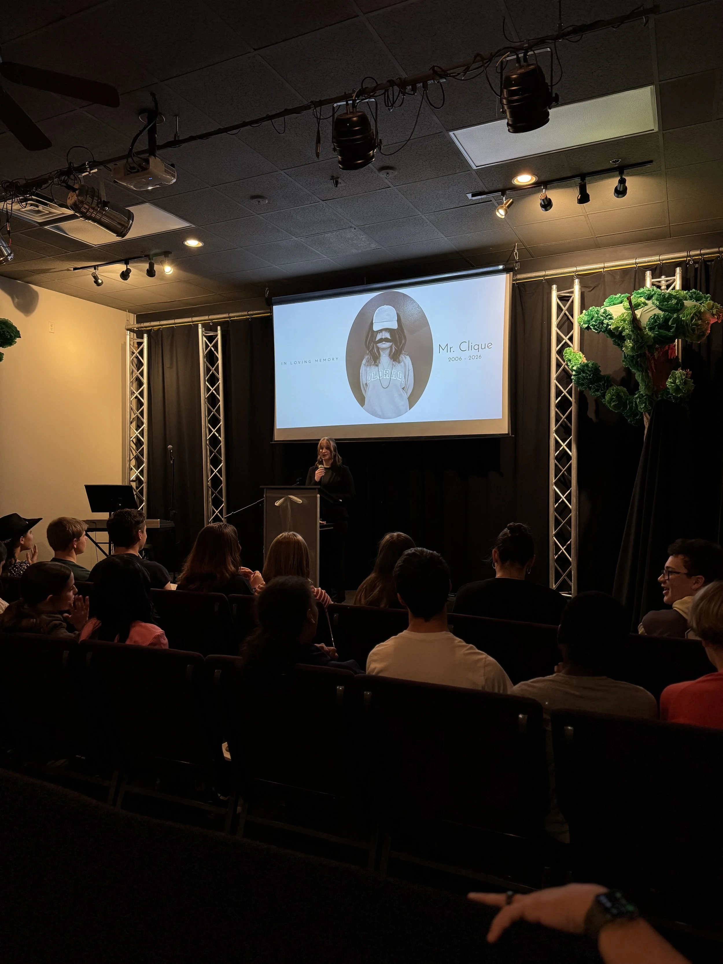 A woman speaking at a podium in front of an audience in a darkened room with a large screen behind her displaying a memorial graphic for Mr. Clique, including a photo of a person with long hair, a baseball cap, and a mustache, with the years 2006-202