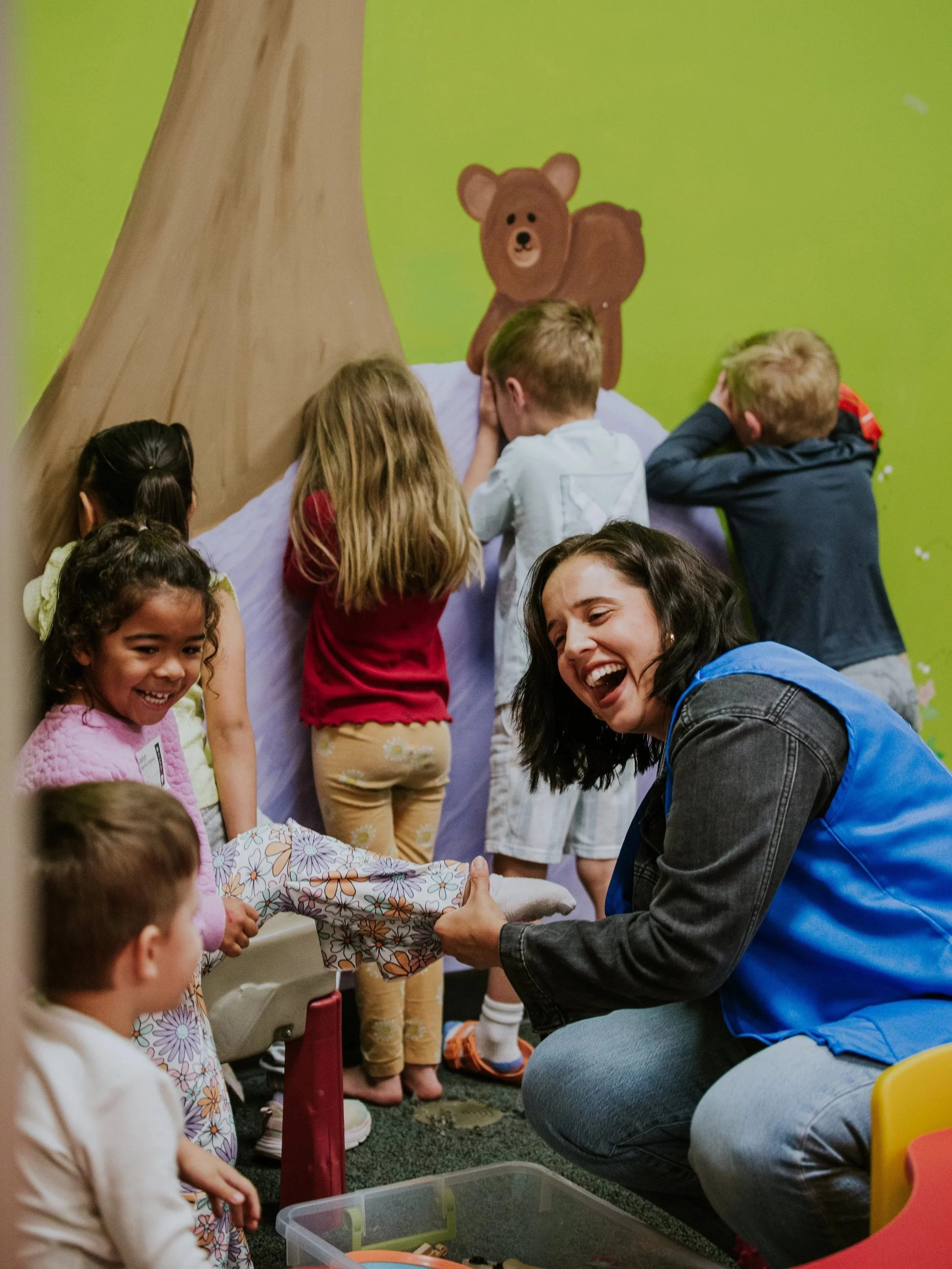 Children and a woman in a classroom participate in a gift exchange or activity. The woman, wearing a blue jacket, is handing a wrapped gift to a girl in a floral dress. The background features a painted mural of a brown bear on a green wall.