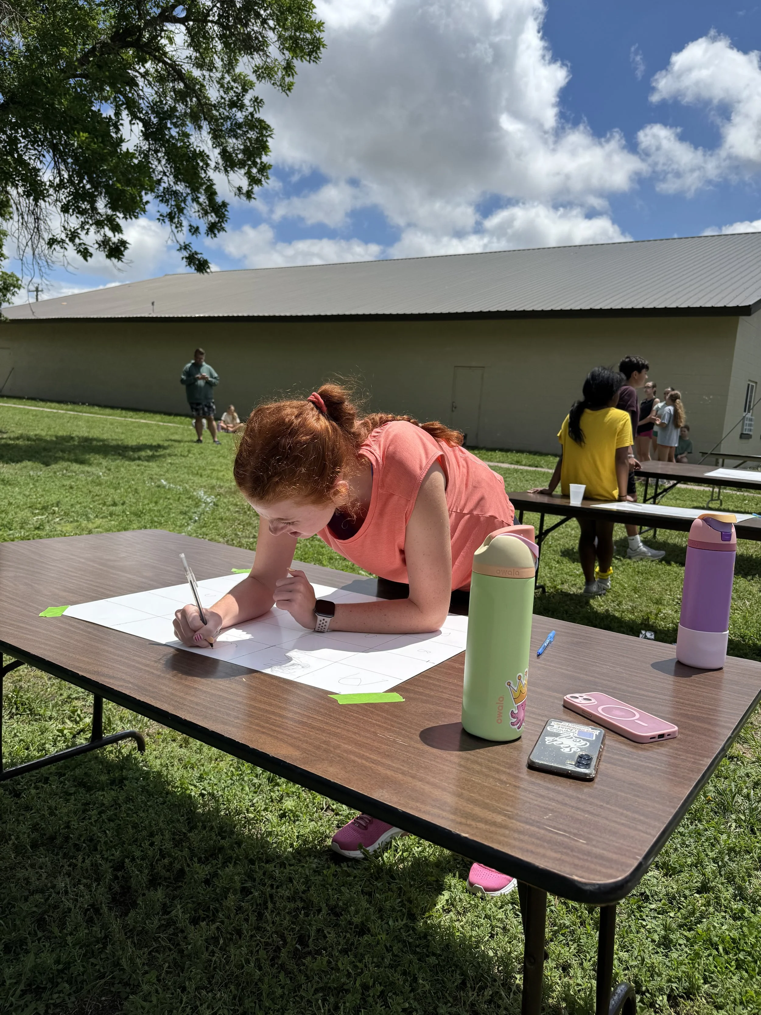 A woman with red hair, wearing a pink shirt, bends over and writes on a large sheet of paper at an outdoor event on a grassy area under a tree. Several people stand and sit in the background, some near a beige building with a sloped roof. On the tabl