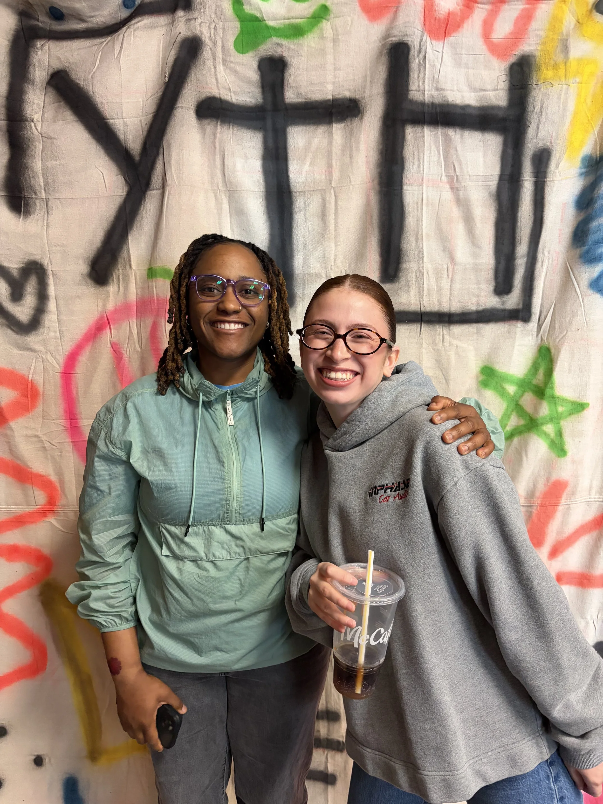Two women smiling, one holding a McDonald's cup with a straw, standing in front of a backdrop painted with graffiti and the words 'YTH' in black spray paint.