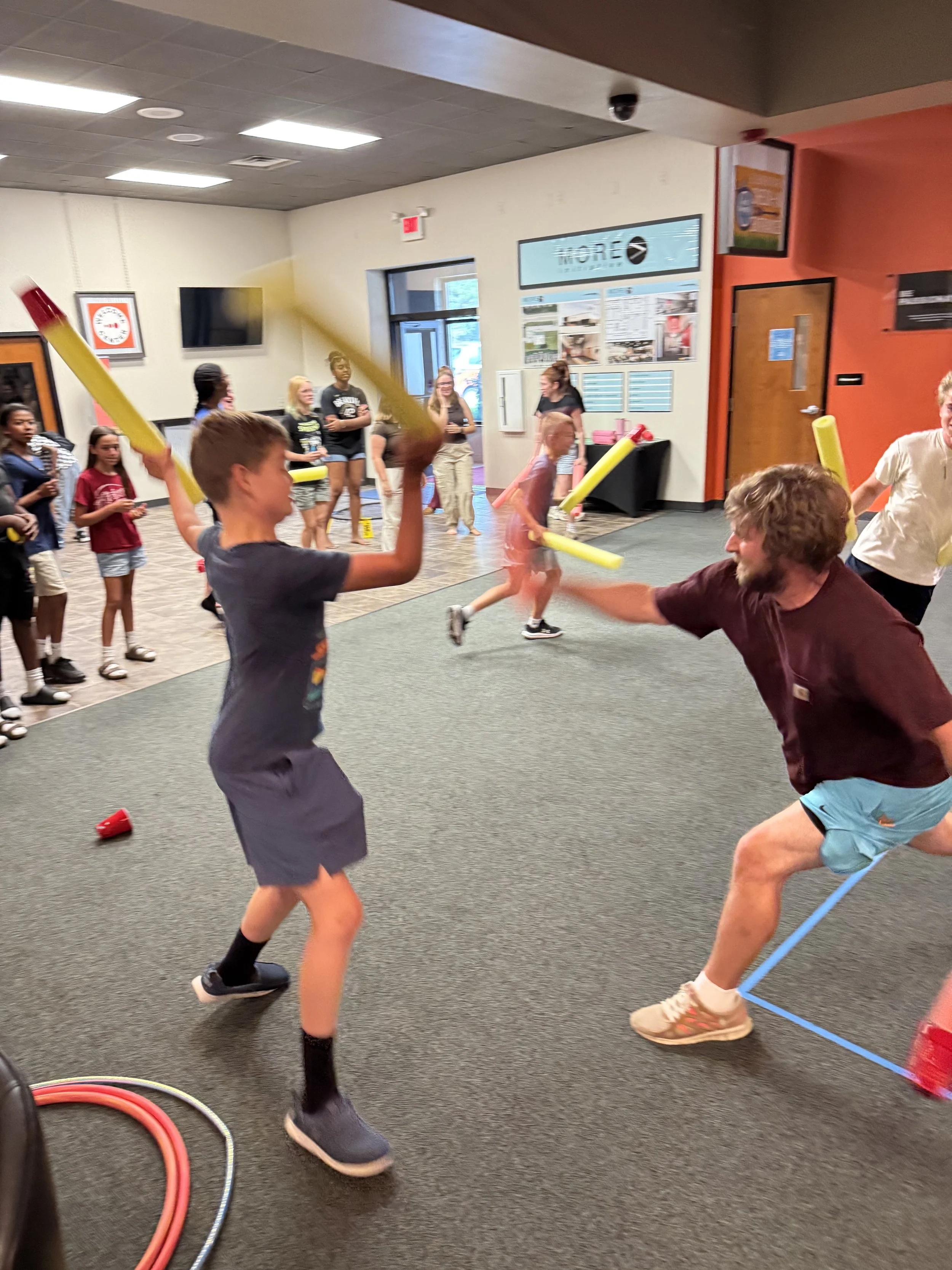 Children and adults playing with foam noodles in an indoor activity area, some are fighting or wrestling, others watch or cheer.