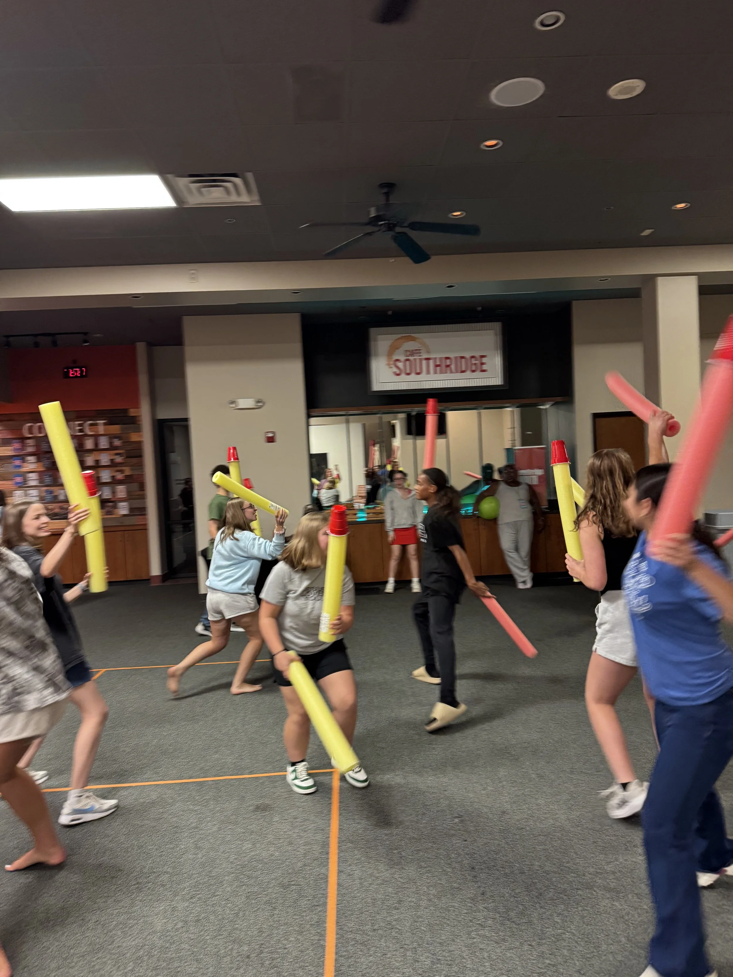 People participating in a foam noodle activity inside a building, with some holding foam noodles and others moving around. The background includes a sign that says 'Southridge' and a group of people observing or participating.