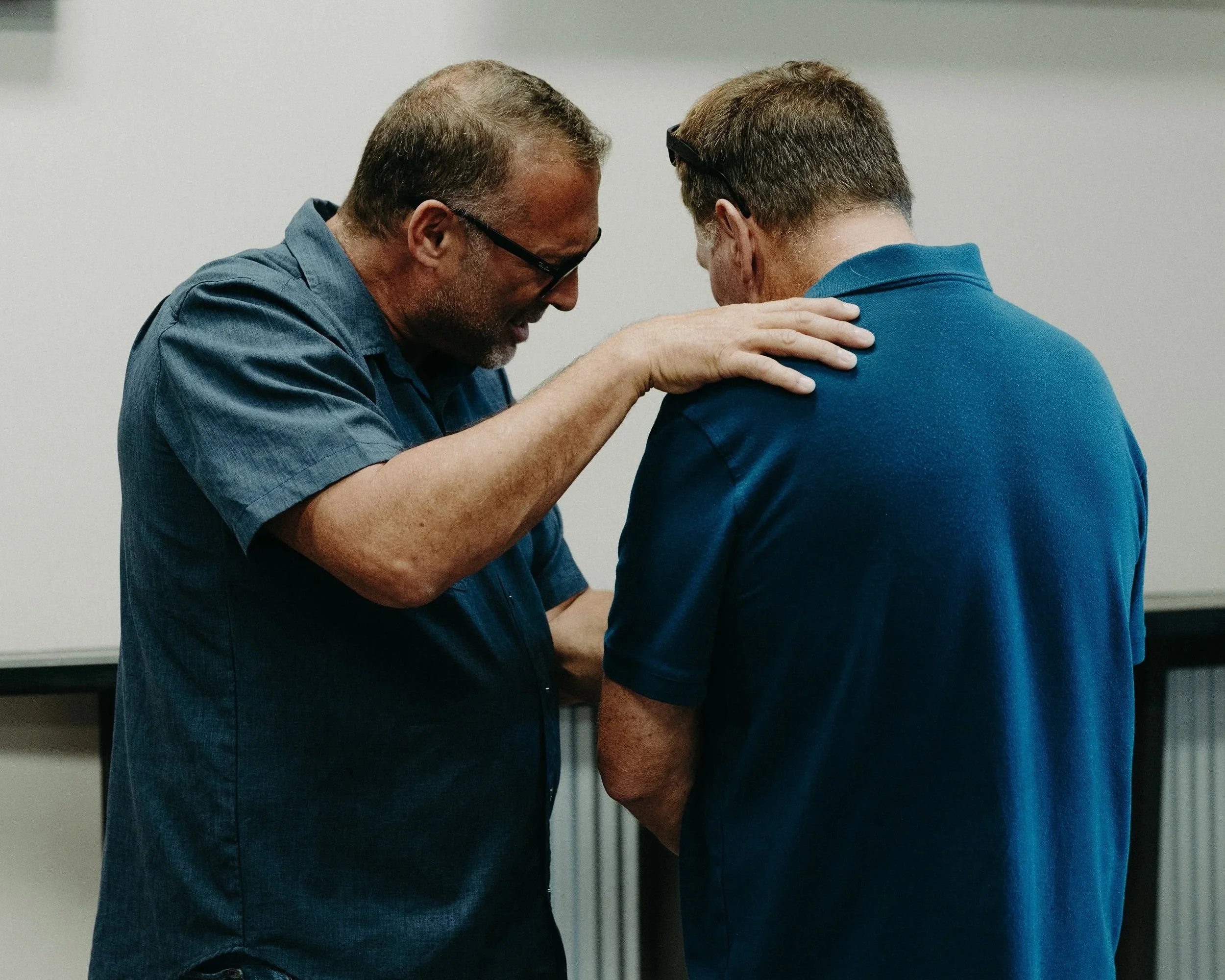 An older man with gray hair and glasses is kneeling, speaking gently with a younger man who has short brown hair and glasses on his head, both wearing blue shirts.