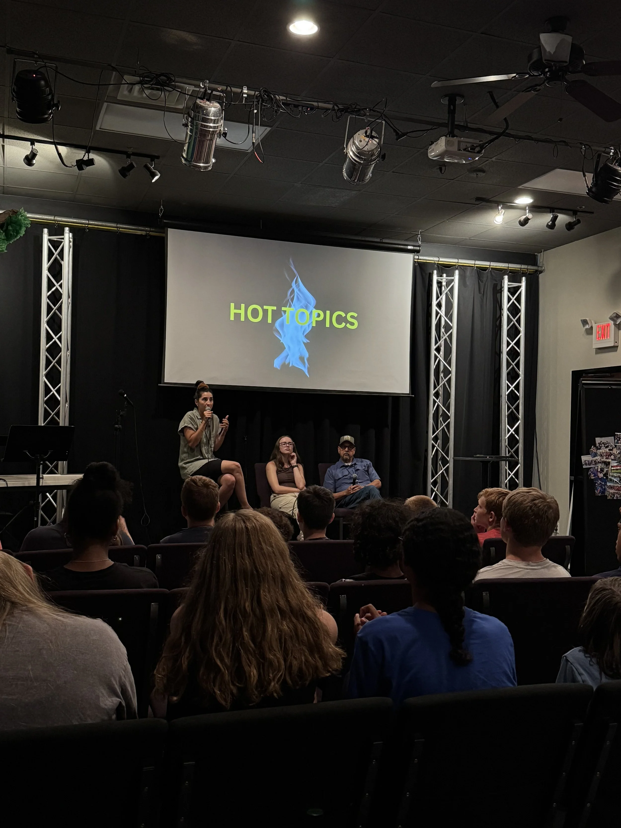 A panel discussion on stage with three people seated, one woman standing and speaking into a microphone, a large screen displaying 'HOT TOPICS' with a blue flame graphic, and an audience watching.