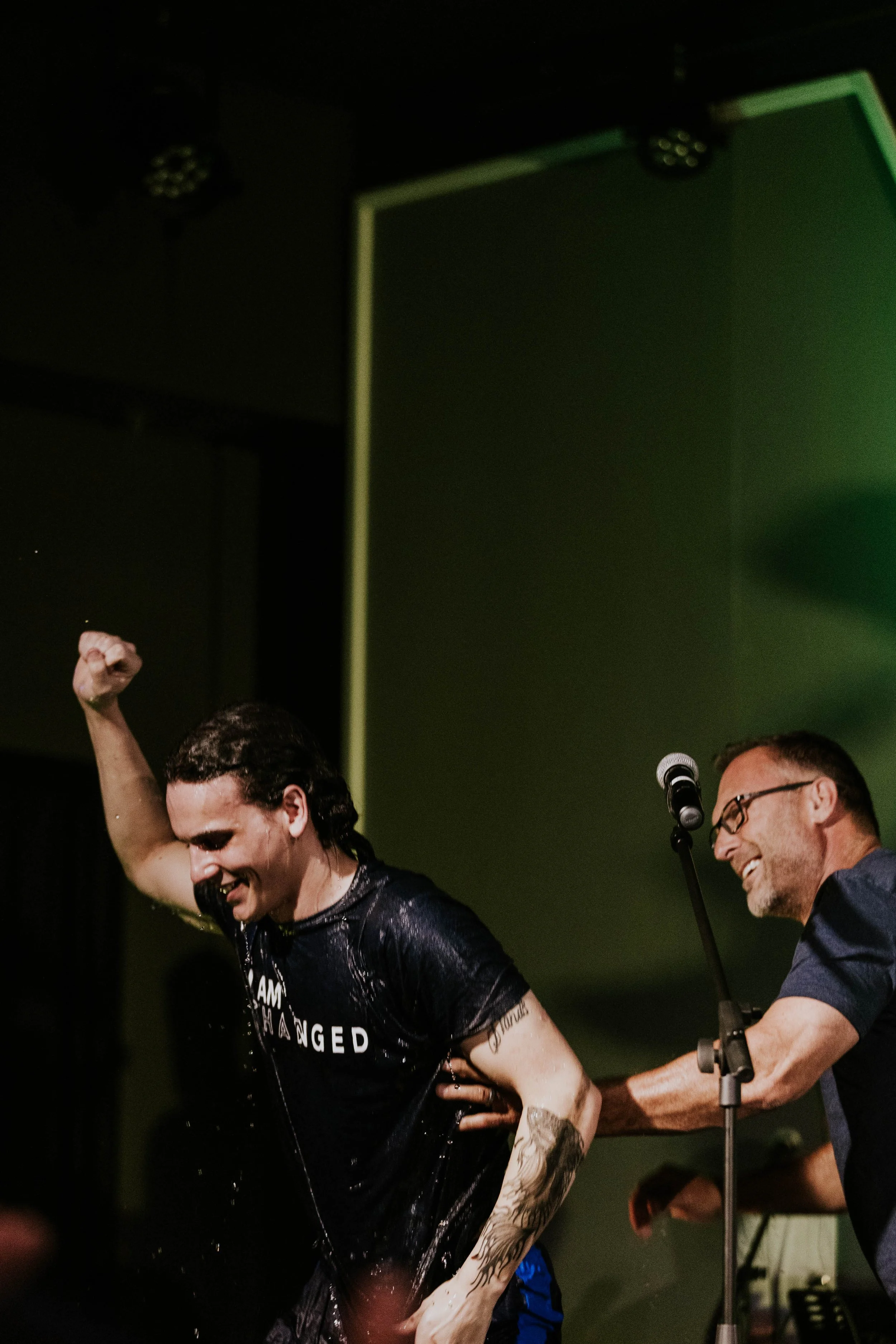 Two people on stage, one with raised fist and wet black shirt, the other smiling near microphone, in a dark room with green lighting.