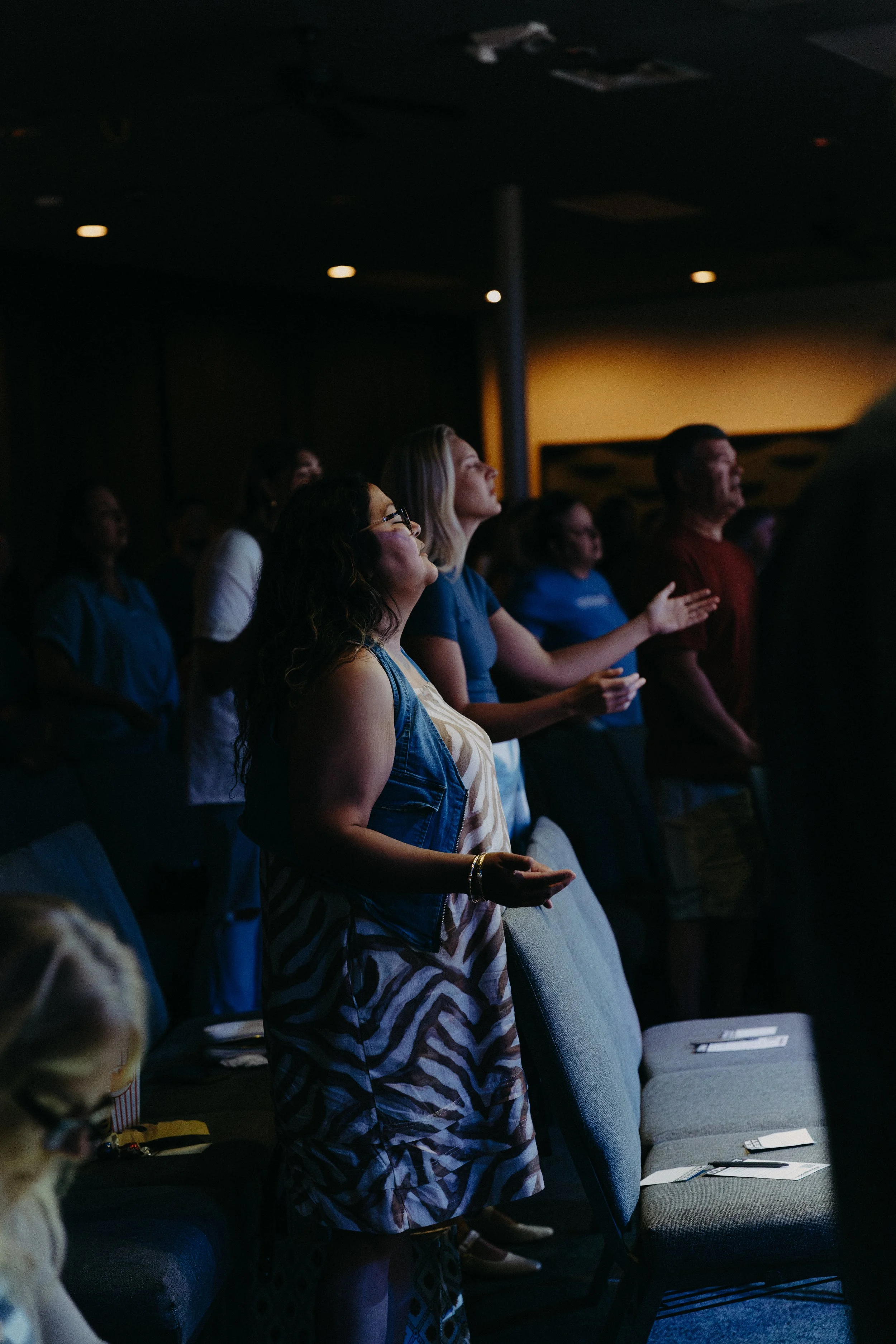Group of people in a dimly lit room, standing with eyes closed, some with hands raised or clasped, attending a spiritual or prayer session.