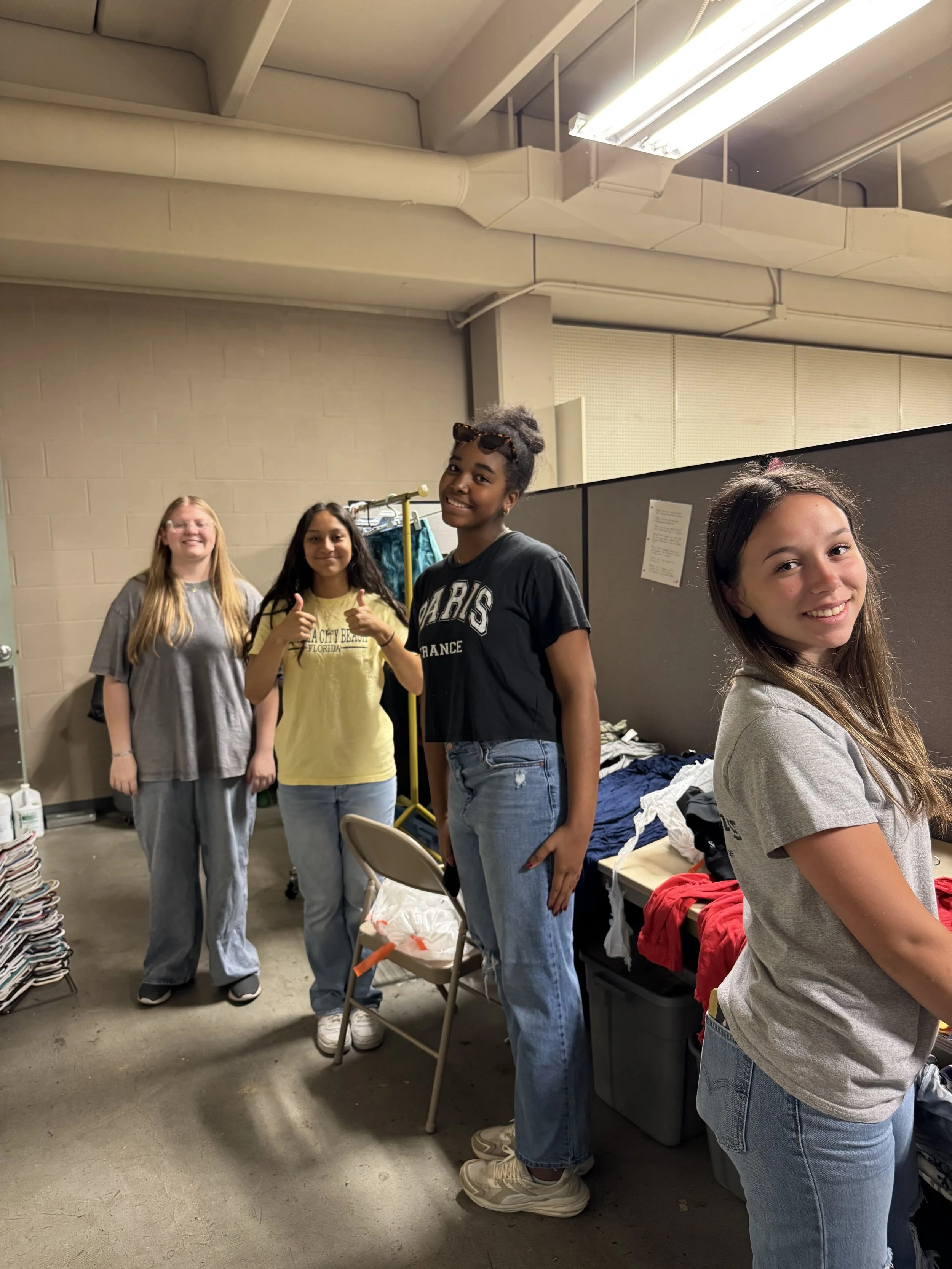 Four young women standing together in an indoor setting that appears to be a storage or backroom area, with piles of clothes and supplies around, smiling and posing for the camera.