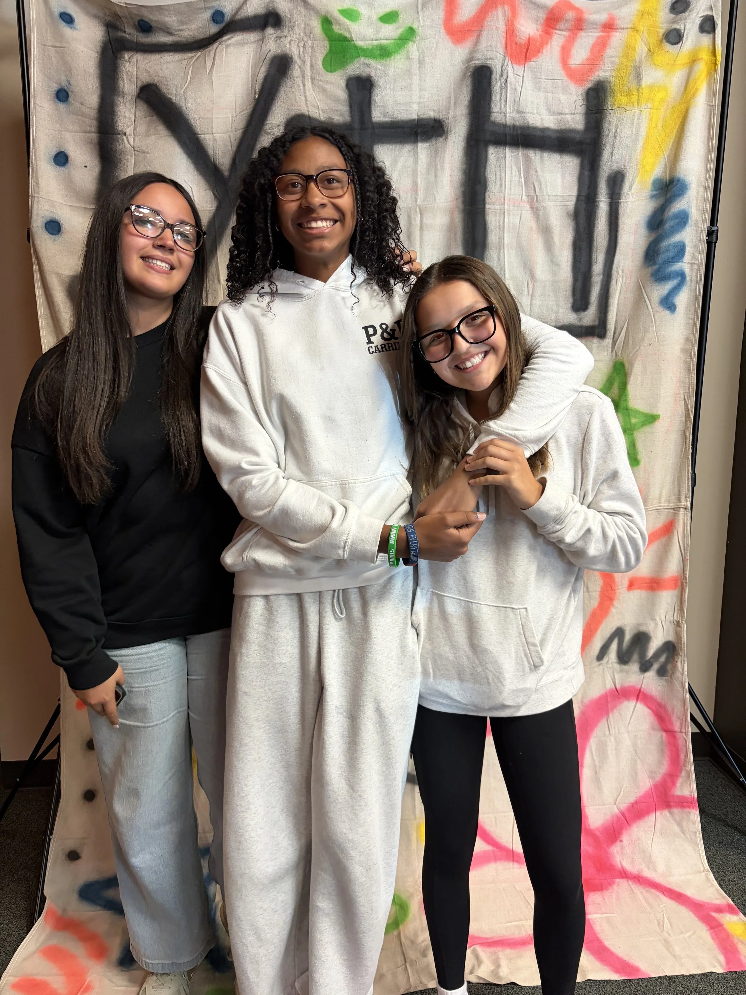 Three young women smiling and posing together in front of a graffiti-style backdrop with colorful spray paint and black letters.