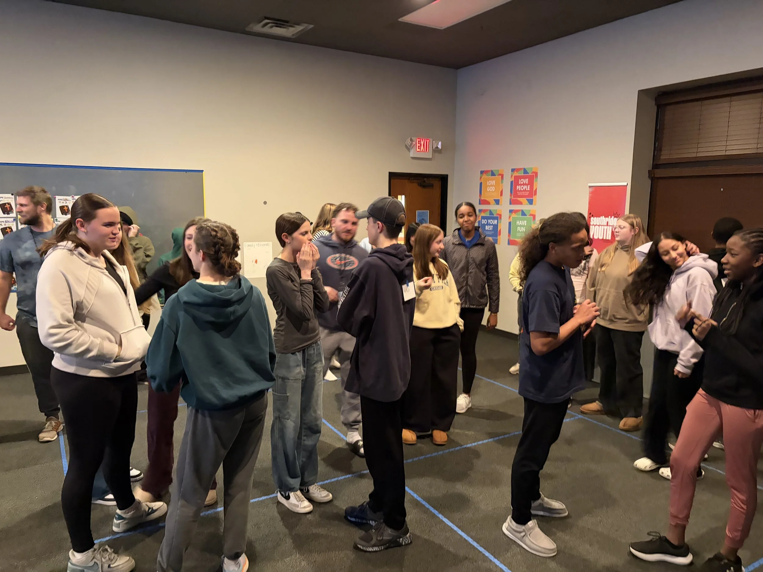 A group of diverse teenagers standing in a room with colorful posters on the wall, engaged in conversation.