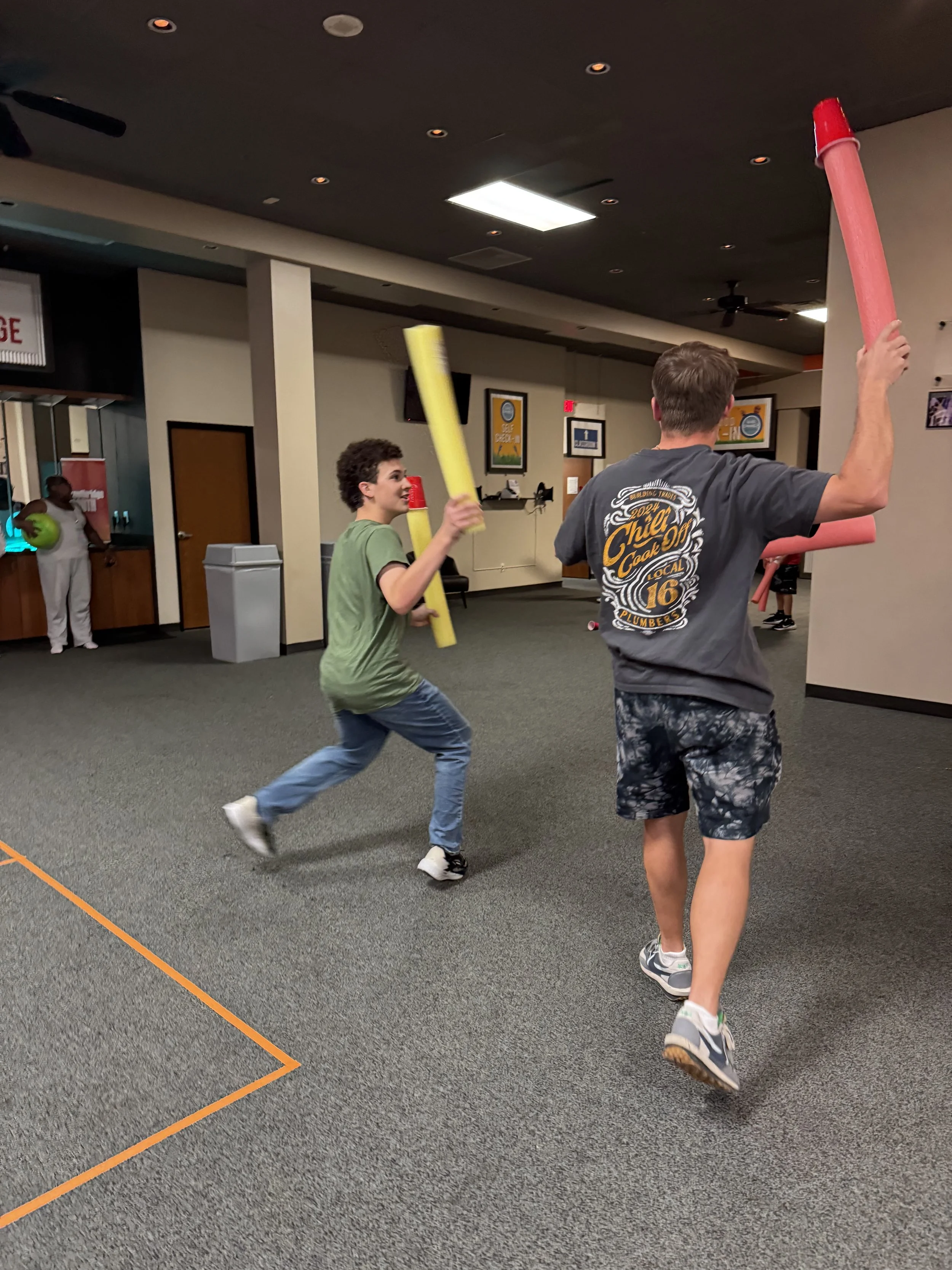 Two young men playing with foam pool noodles inside a room with carpeted floor, while a third person holding a green ball stands near the back wall.