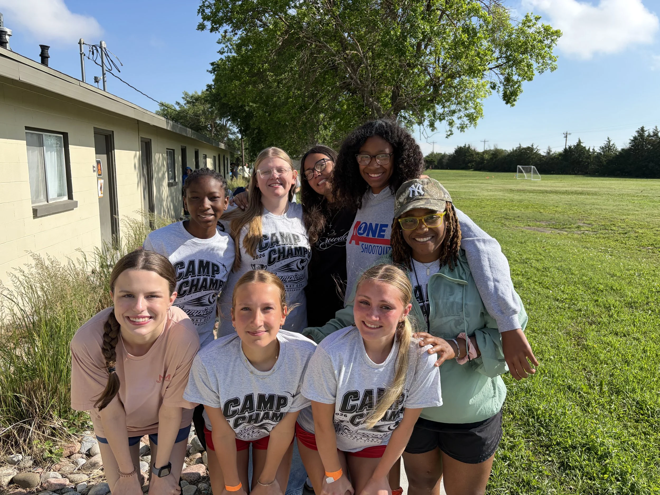 Group of smiling young girls and women outdoors, some wearing camp shirts, with a building, tree, and grassy field with soccer goals in the background.