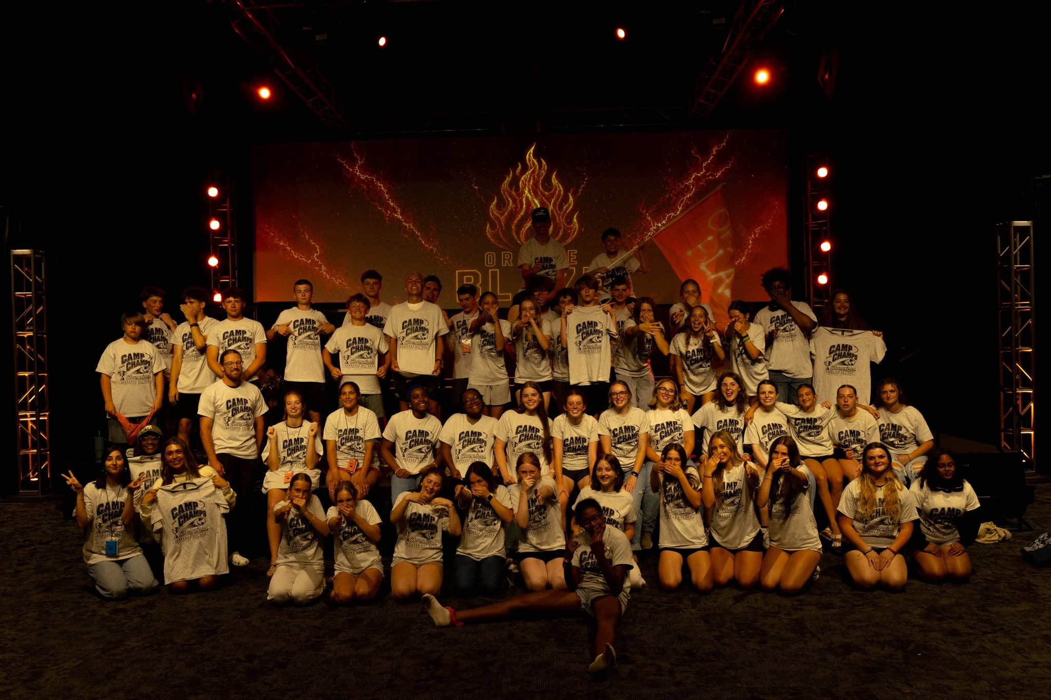 Group of young people on stage at a camp event, wearing white t-shirts with 'Camp Champ' printed on them, with a large screen in the background displaying lightning-like graphics and the words 'Orange BLAST'.