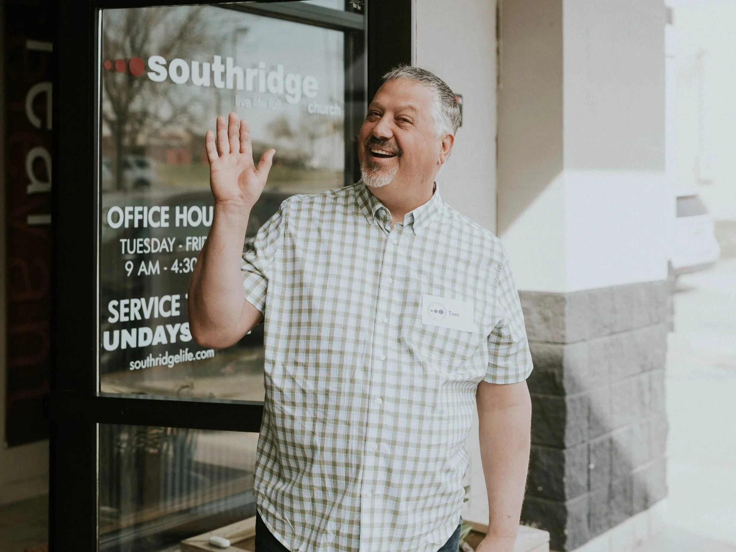 Smiling man in checkered shirt waving at the camera, standing in front of the church office window with hours listed, outside in a bright, open area.