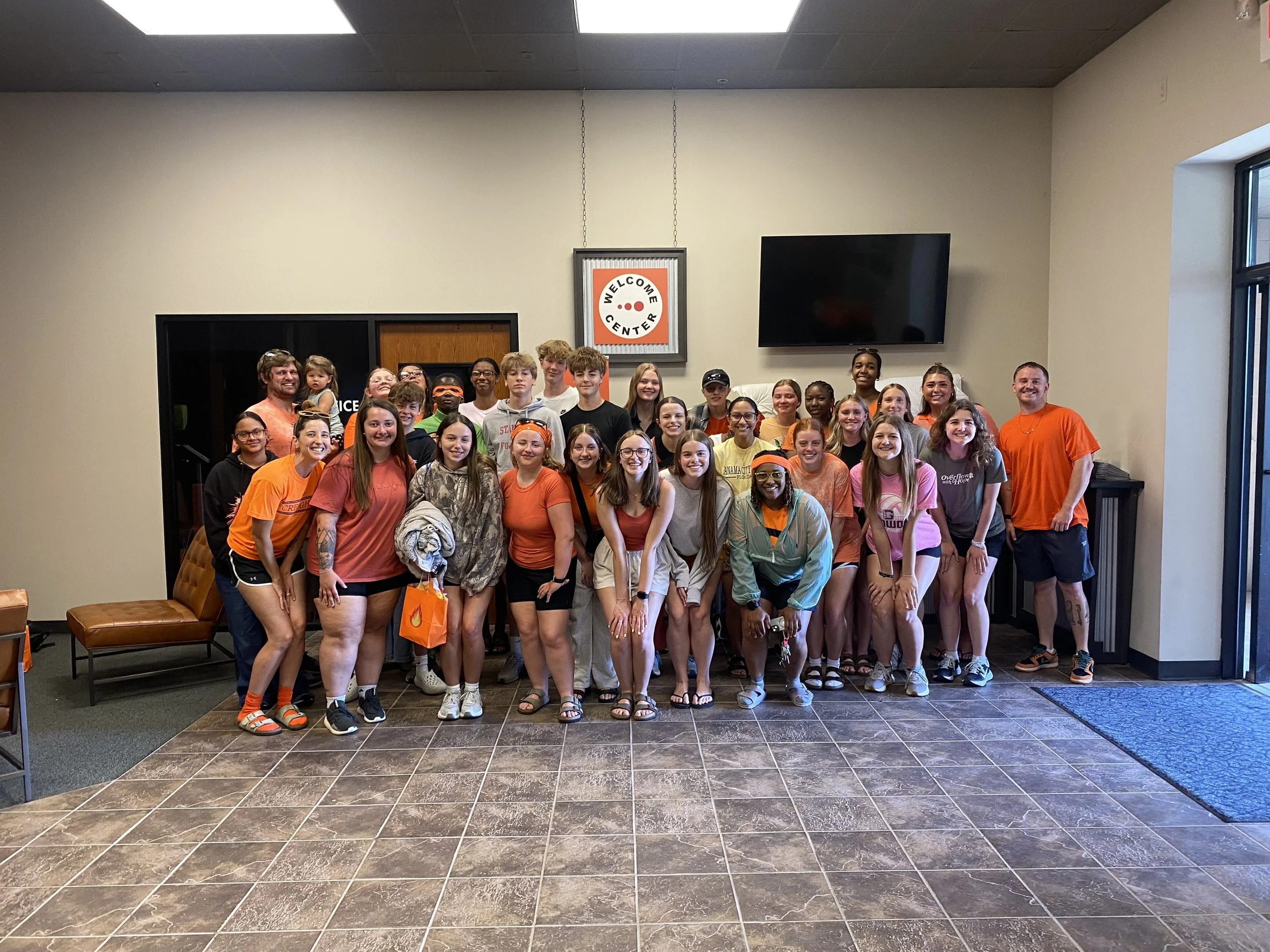 A large group of diverse people of different ages and genders smiling and posing in a building lobby with a 'Welcome Center' sign. Everyone is dressed casually, with some wearing bright colors, and standing close together.