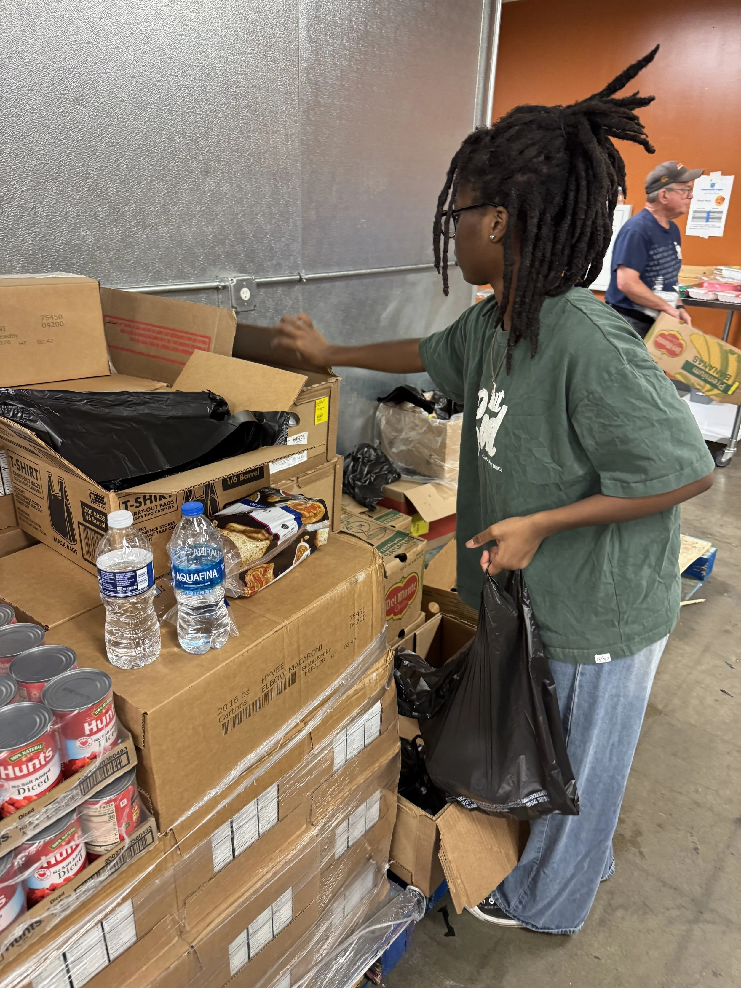 A person with dreadlocks wearing glasses and a green T-shirt is shopping at a food bank or food pantry, selecting canned and boxed food items packed on cardboard boxes and shelves.