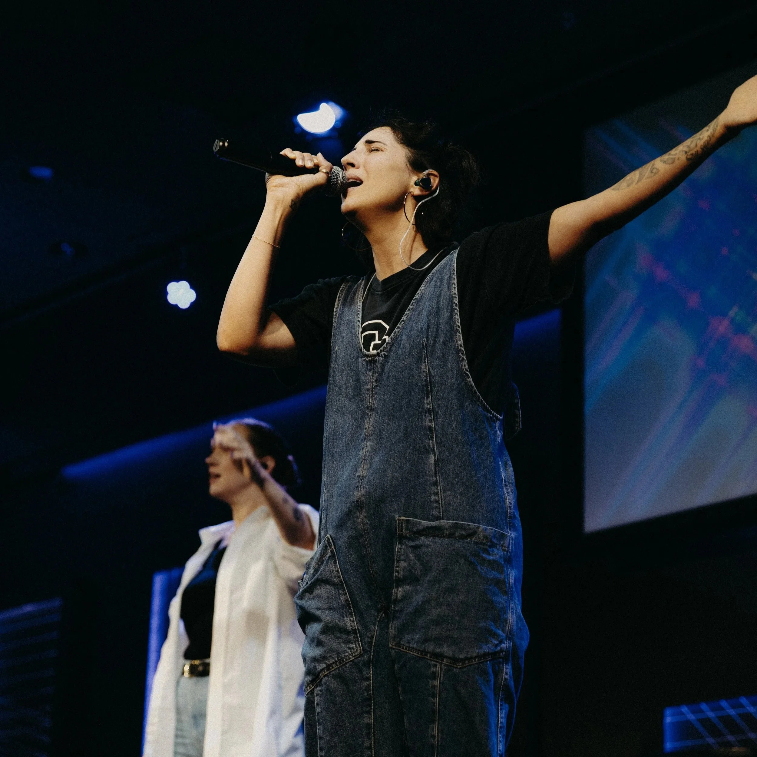A female performer singing into a microphone on stage, with her arm raised, tattoos visible on her arm, wearing hoop earrings, a black t-shirt, and denim vest, with a dark background and stage lighting.