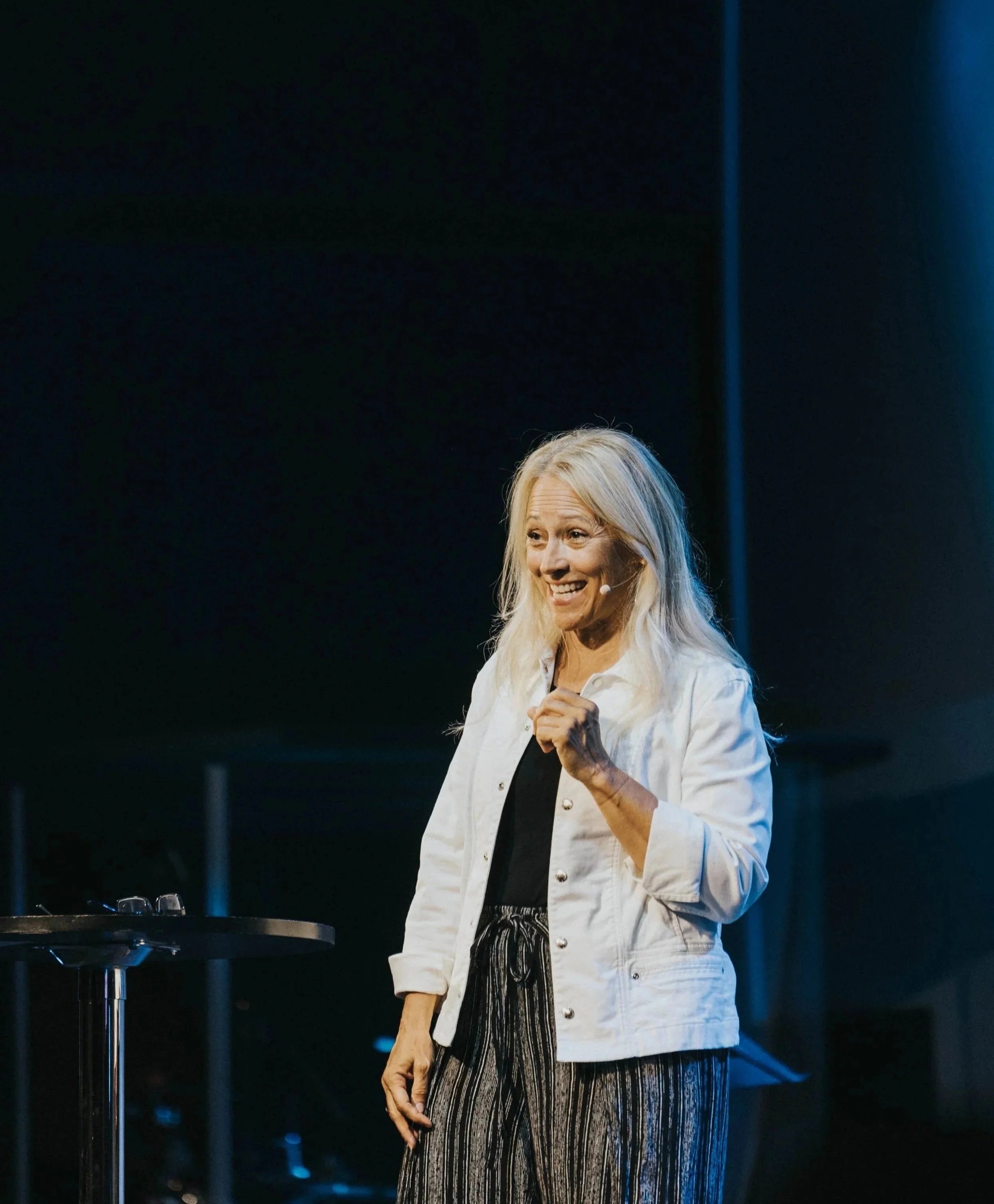 A woman with long blonde hair standing on stage, smiling, wearing a white jacket and black and white striped pants, with a dark background and a small table with glasses on it nearby.