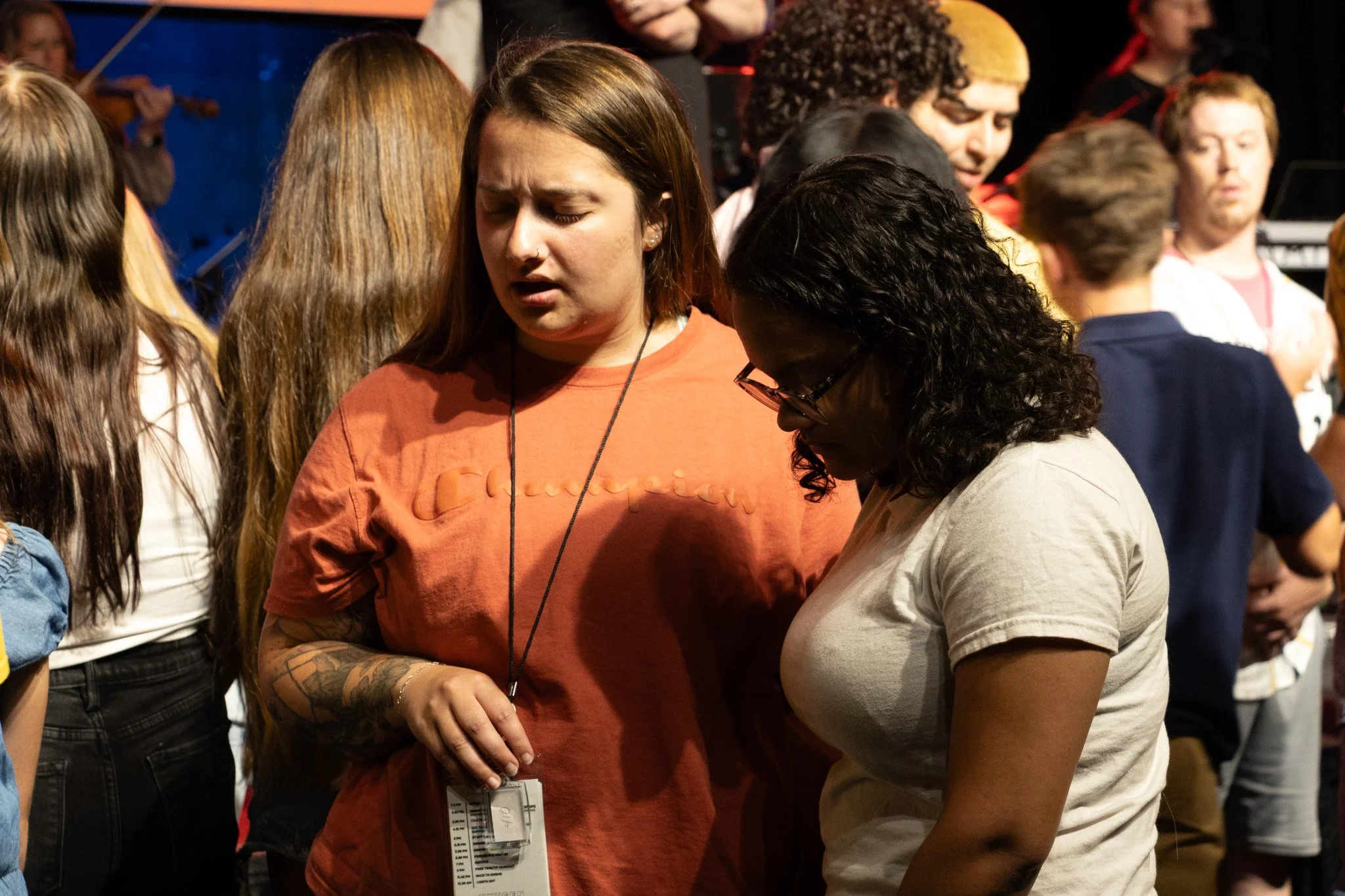 Two women are standing close together, focusing on something in front of them. One has long, light brown hair and is wearing an orange shirt with a lanyard and ID badge. The other has dark, curly hair, glasses, and is wearing a beige shirt. They are 