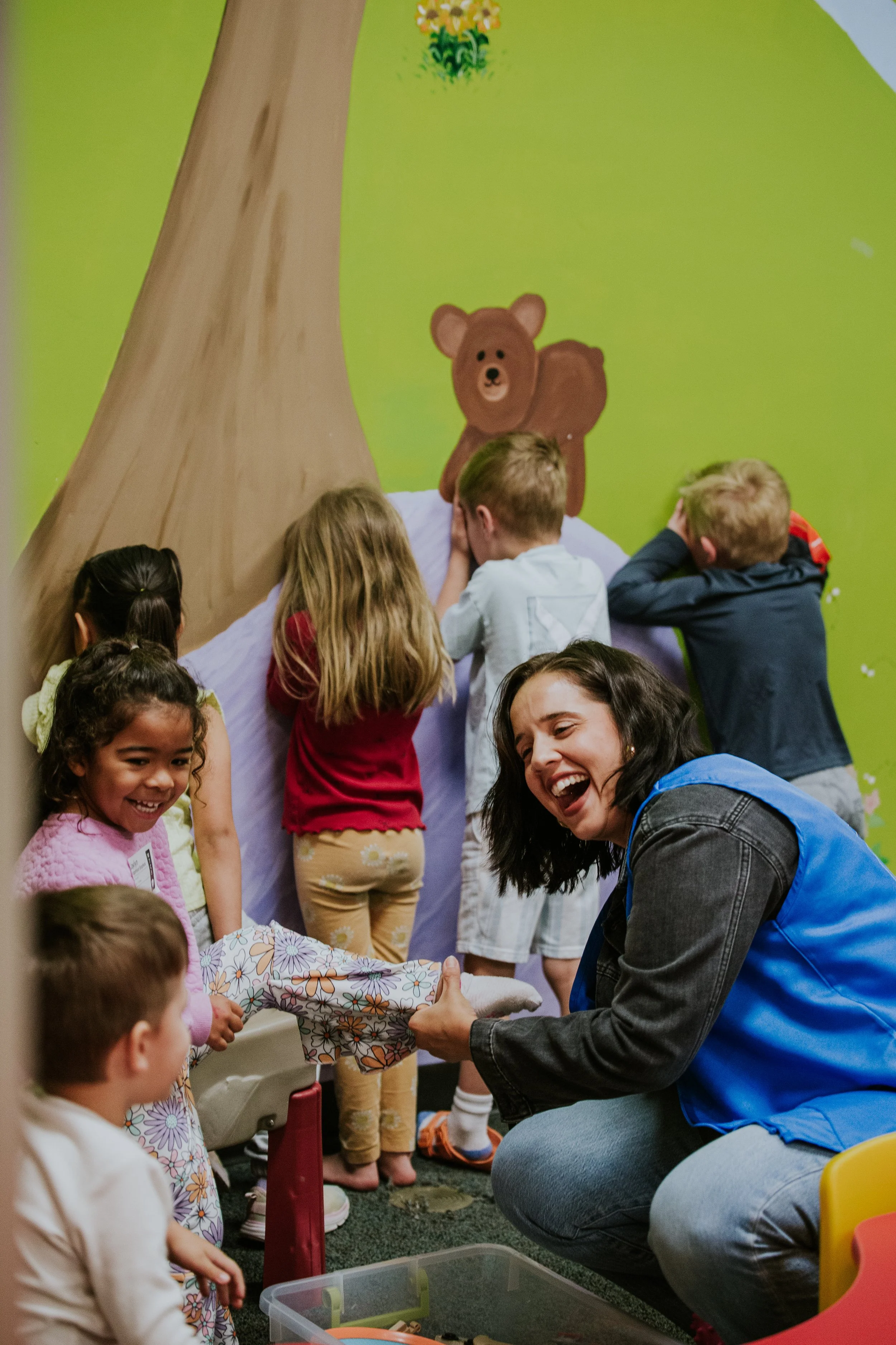 Children gathered around a woman who is helping them with a gift, in a room with a colorful wall featuring a bear and trees painted on it.