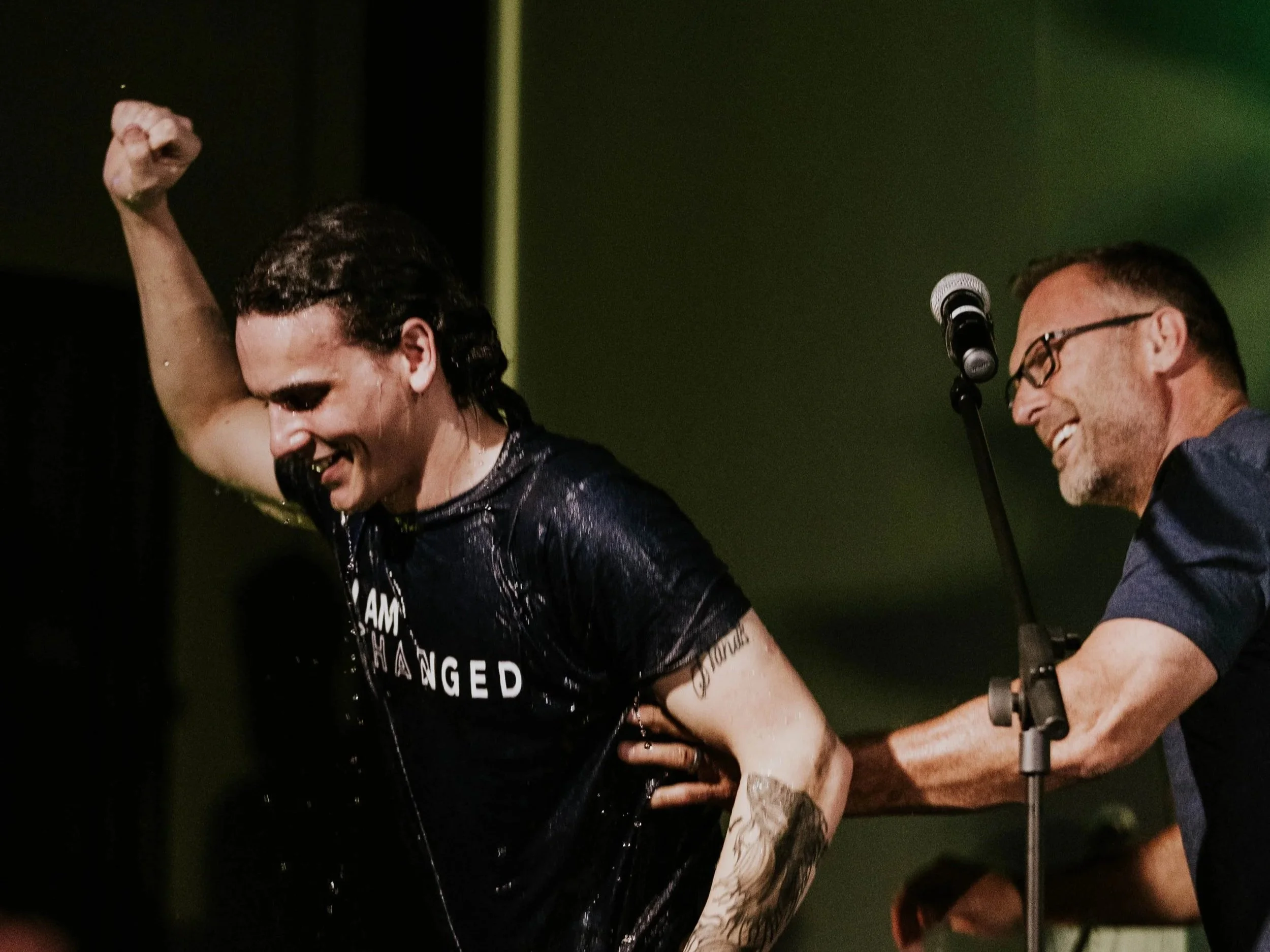 Two men on a stage, one with long dark hair, wearing a black shirt that reads 'I Am Changed,' raising his arm and smiling, while the other with short hair and glasses, standing behind a microphone, also smiling.