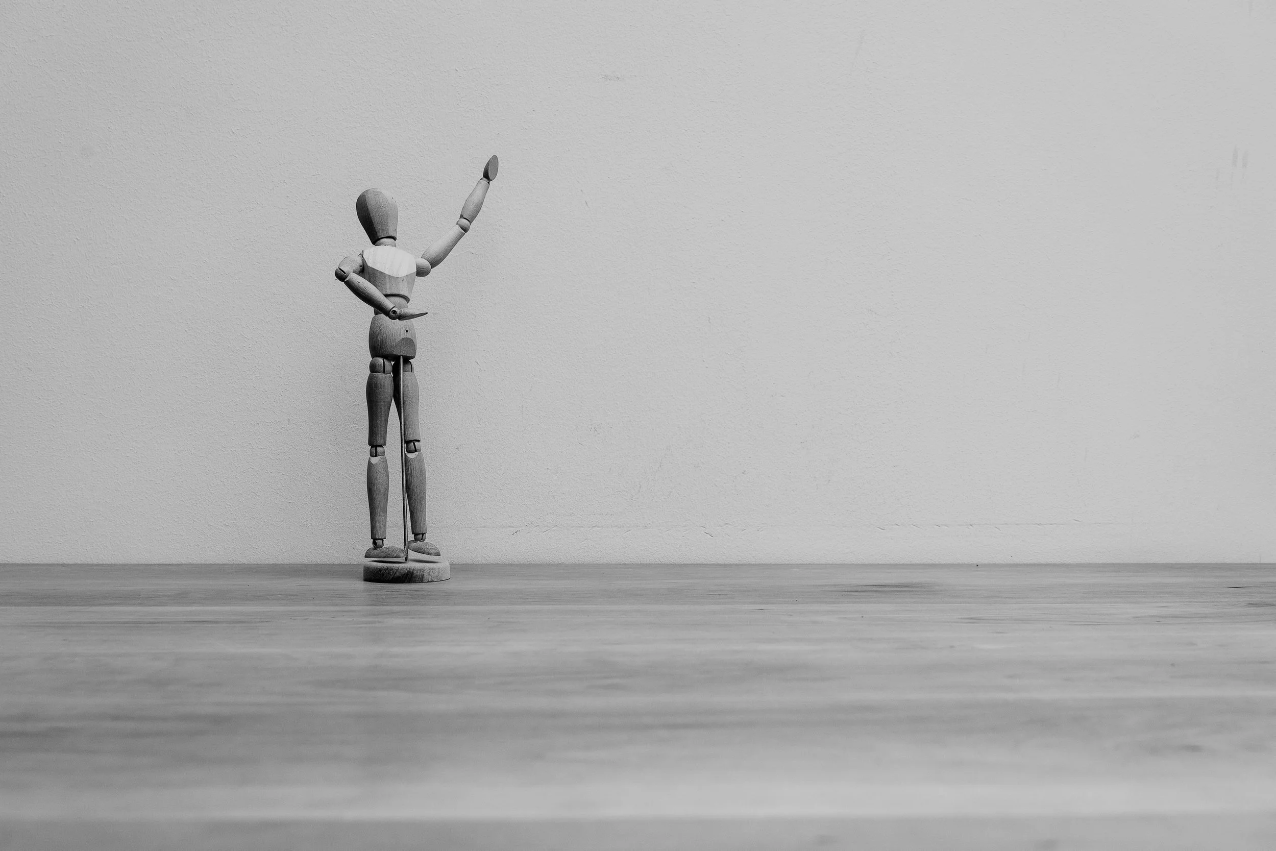 A wooden artist mannequin standing on a circular base on a wooden surface, raising its right arm in the air, against a plain textured wall background.