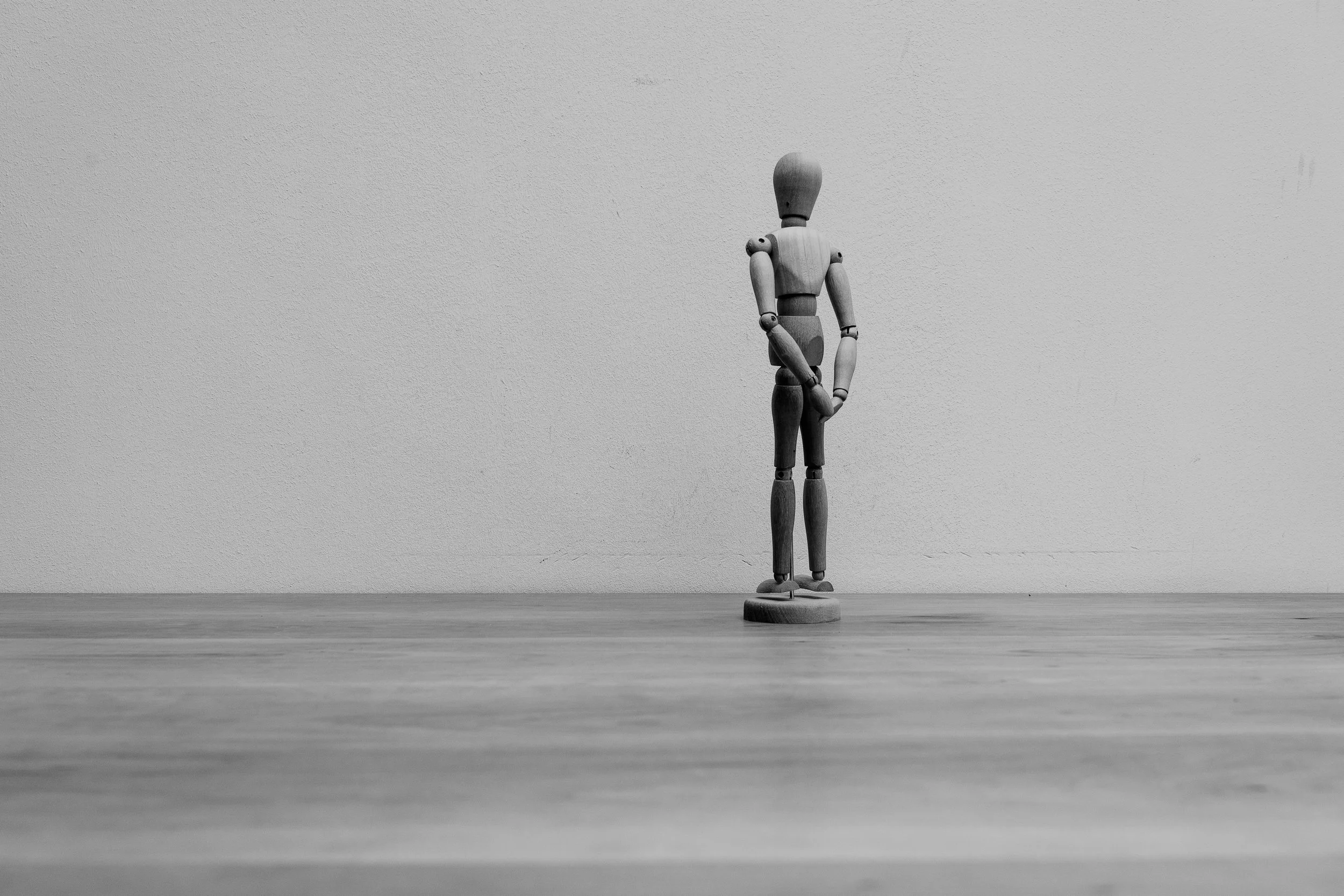A wooden artist mannequin standing on a round base on a wooden surface, posing with one arm bent and one leg crossed, against a plain textured wall.