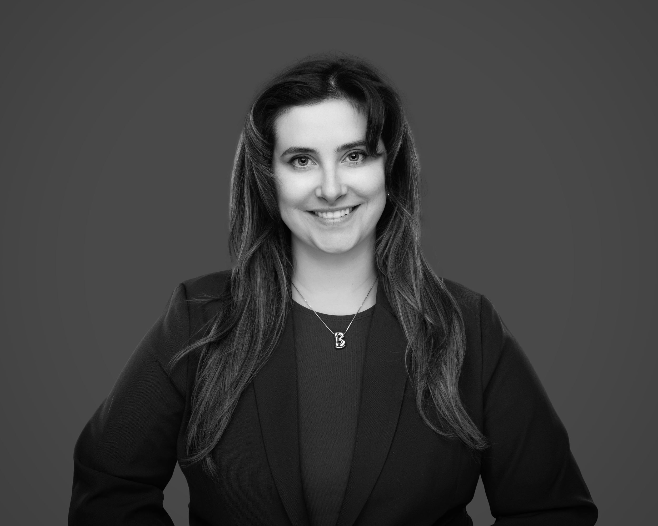 Black and white portrait of a woman with long hair, wearing a dark blazer and a necklace with a letter 'B', smiling against a plain background.