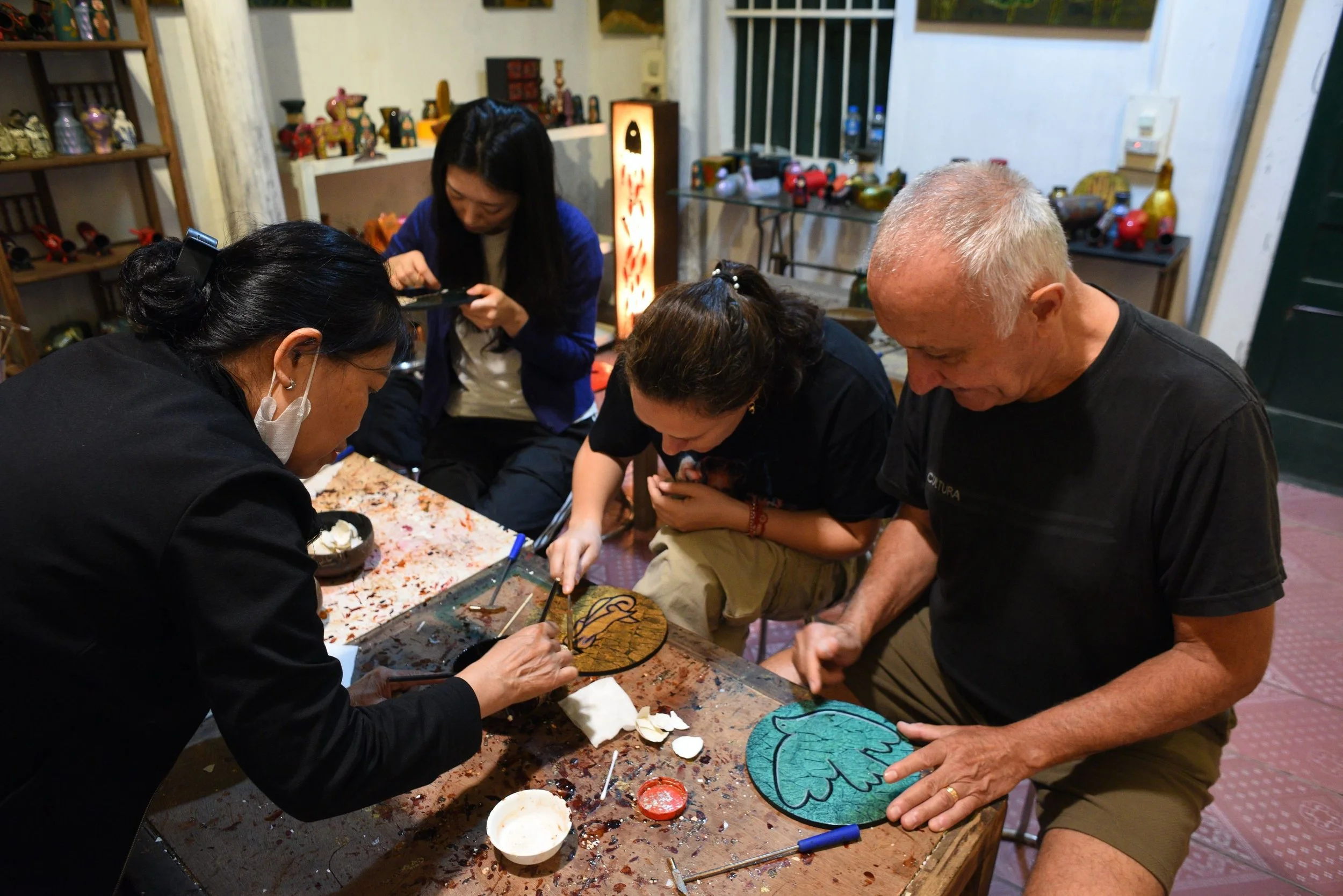 People gathered around a table sculpting and decorating a large wooden leaf. The background features shelves with ceramic and wooden art pieces, and a tall lighted lamp with red and black accents.