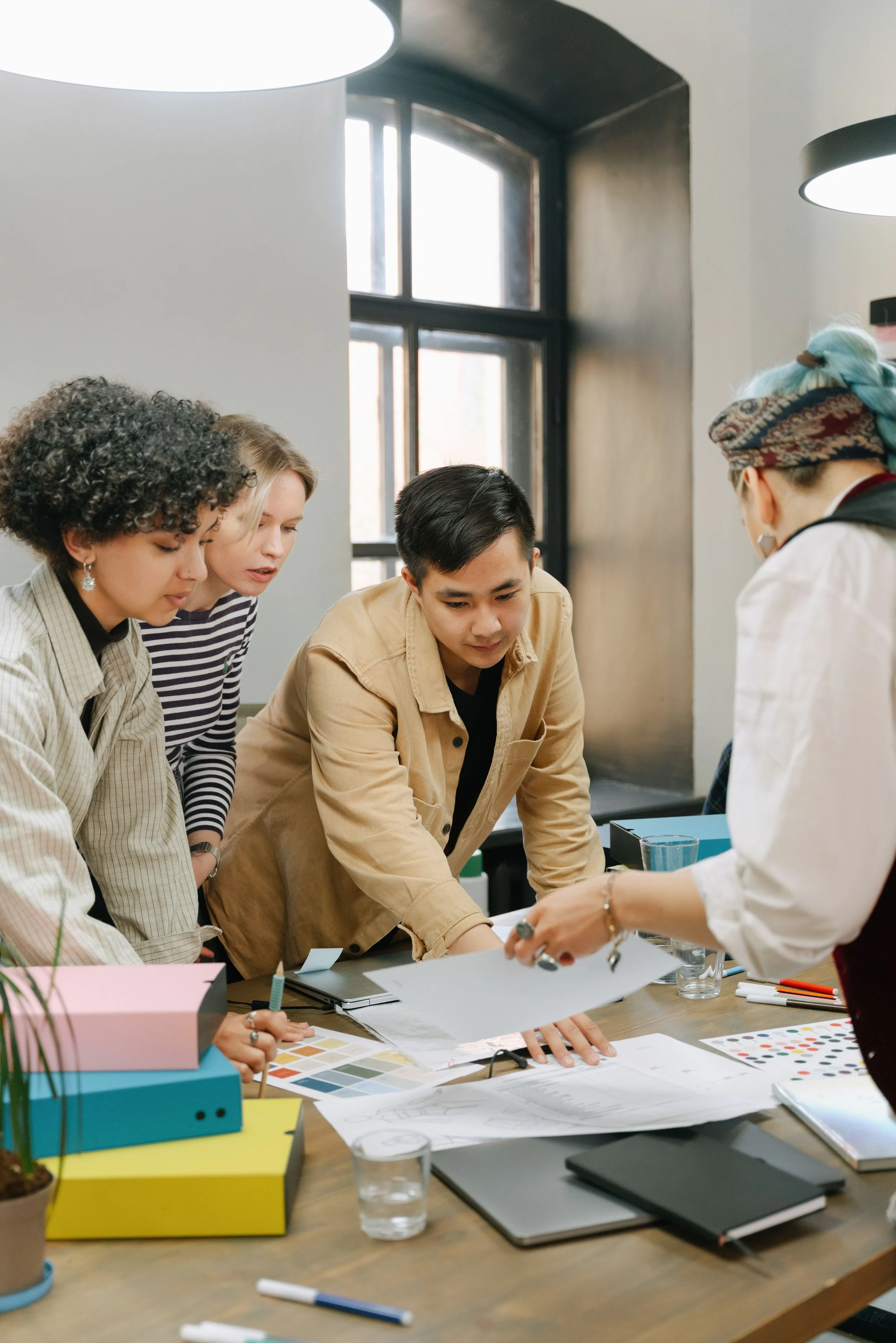 Five people gathered around a table in a bright room, examining documents, color swatches, and design samples, with stationery and notebooks on the table.
