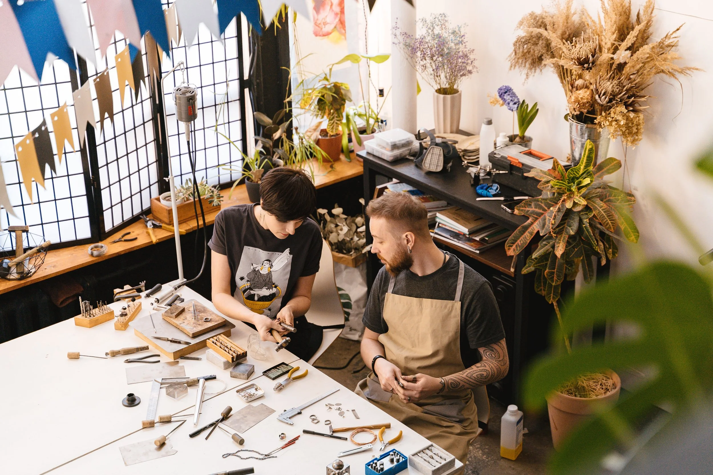 Two people working together at a workspace, surrounded by various tools and jewelry pieces, with potted plants and floral arrangements in the background.
