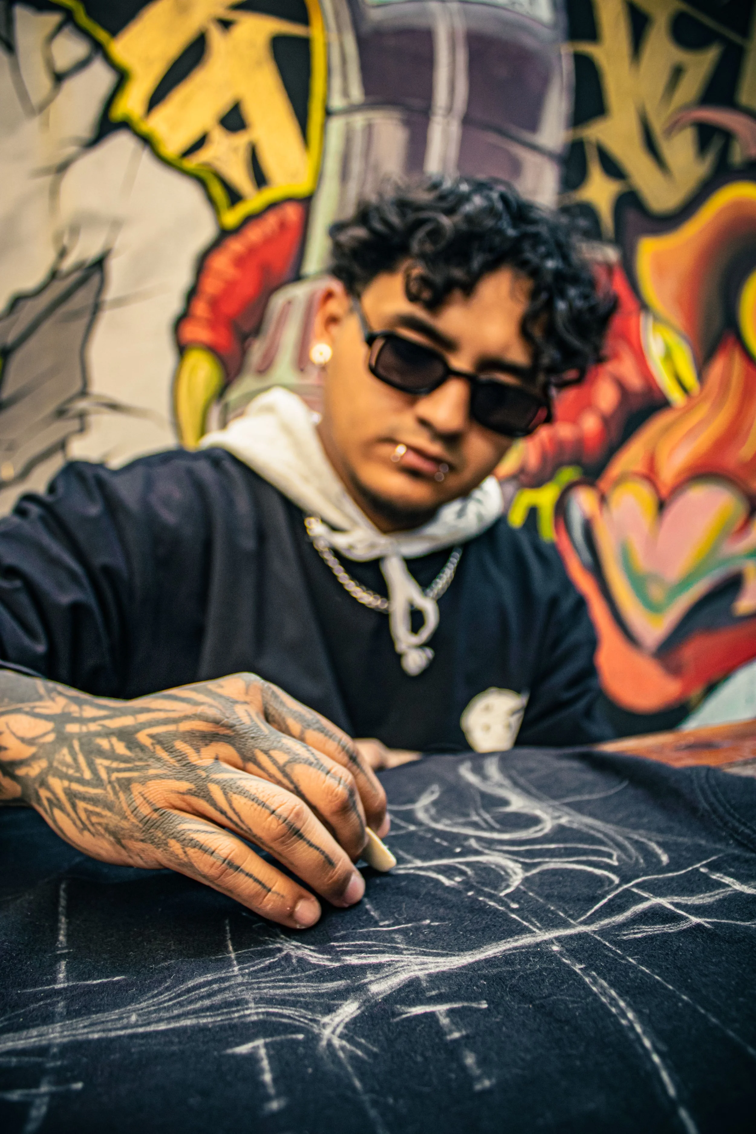 A person with tattoos on their hand drawing on a black surface with a white chalk or crayon, in front of a colorful graffiti wall.