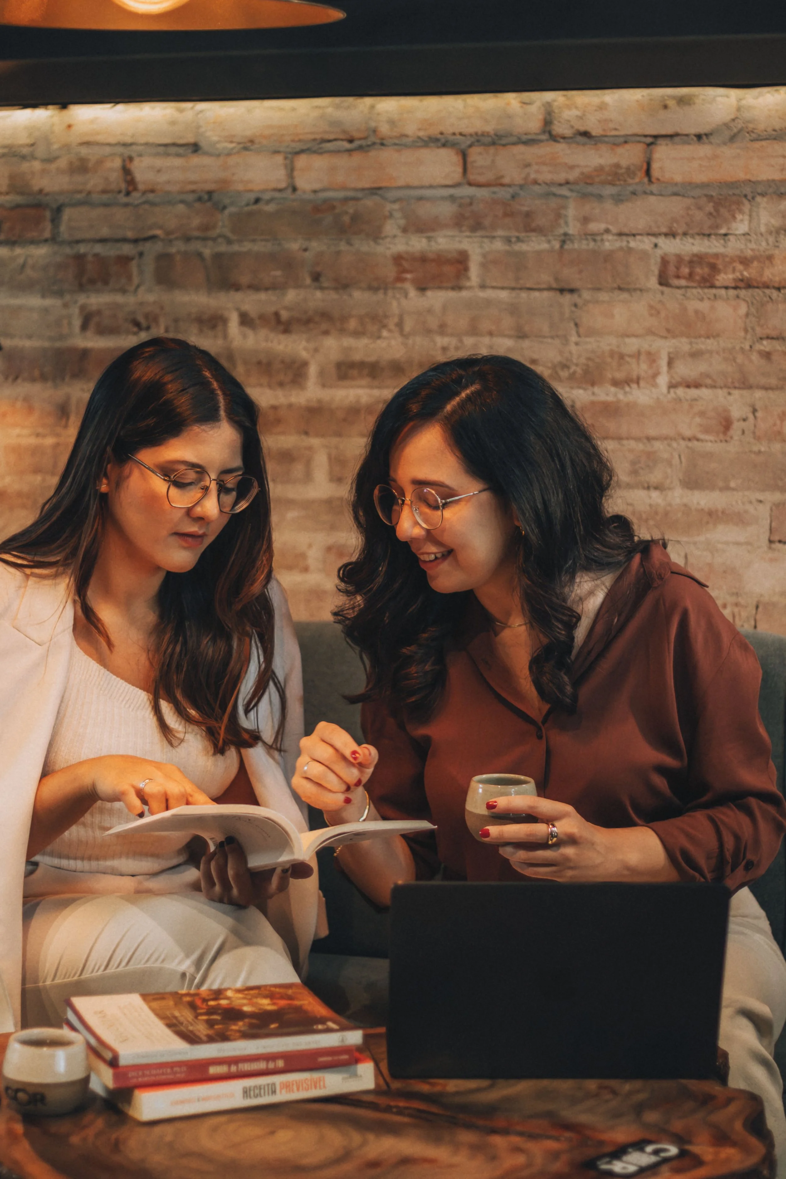 Two women sitting together at a table in a cozy, brick-walled cafe, looking at a menu and smiling, with a laptop and books on the table.