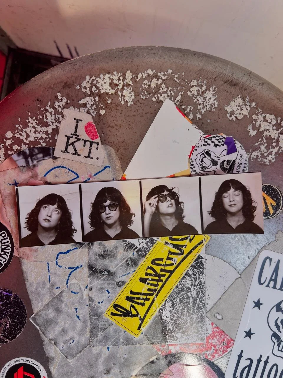 A strip of four black-and-white photo booth pictures of a woman with curly hair, showing different expressions and gestures, placed on a dirty, chipped metallic surface with various stickers and torn paper around.