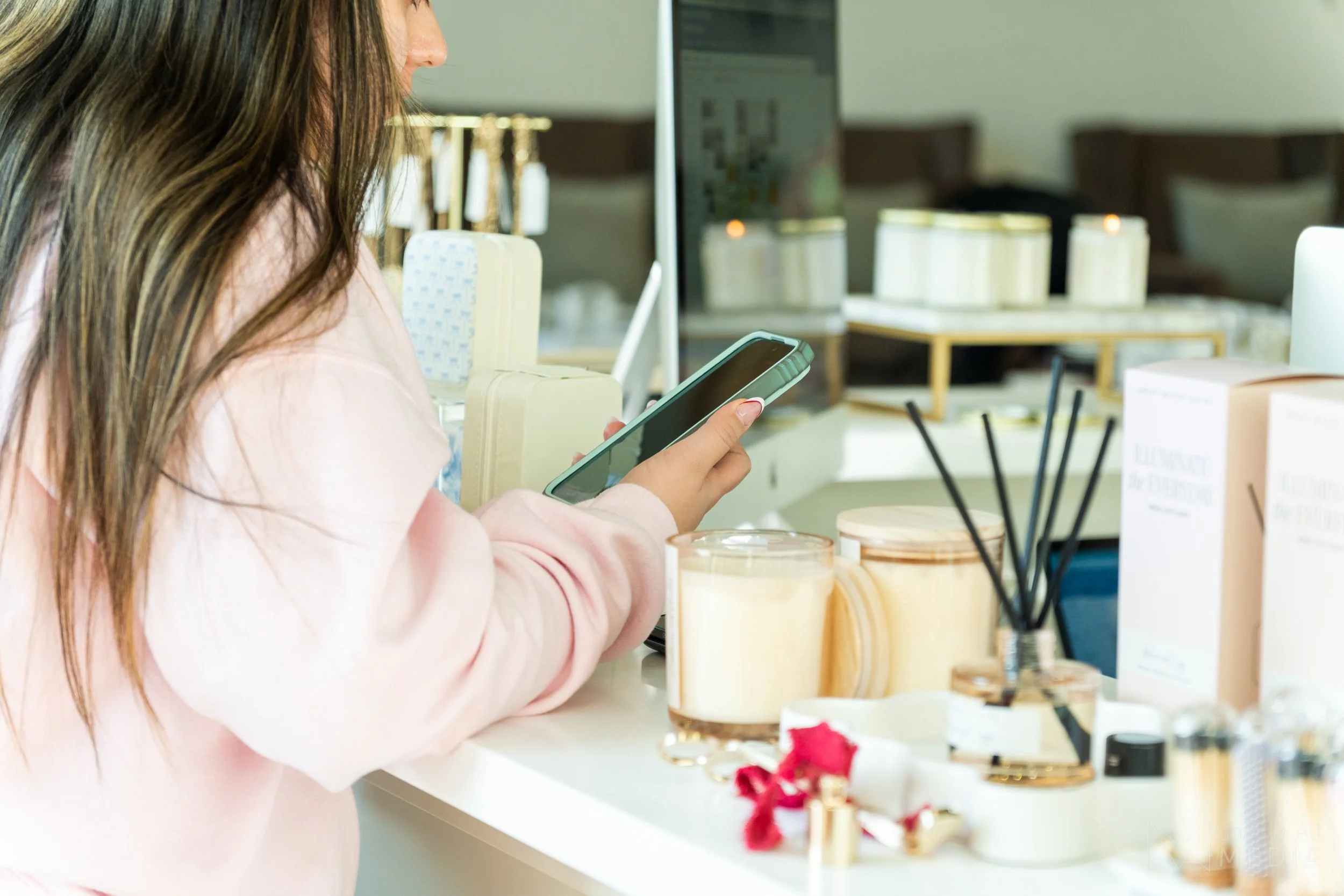 A woman with long brown hair looking at her smartphone in a store, surrounded by candles and beauty products on display.