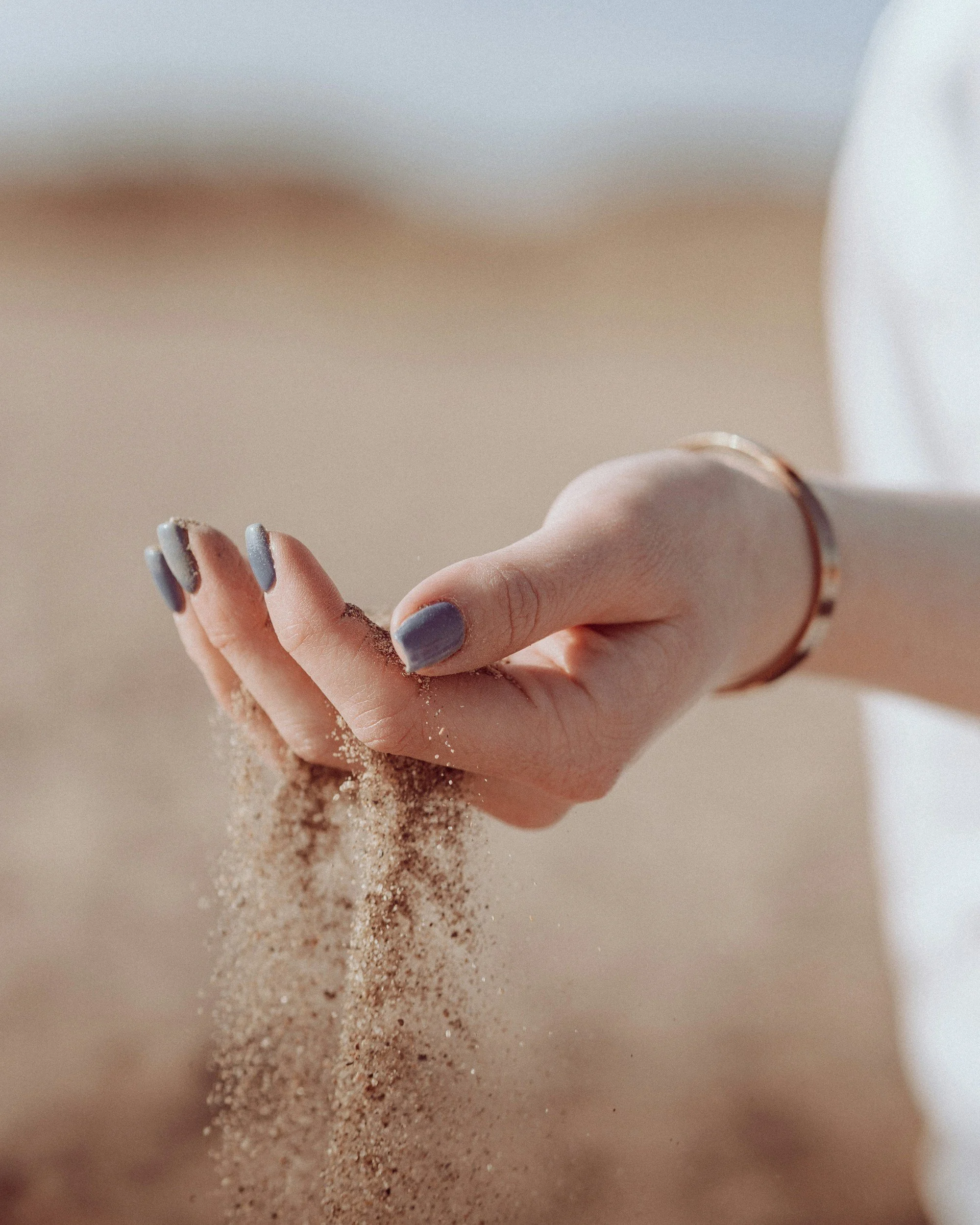 Close-up of a hand with light blue nail polish, holding a handful of fine sandy material, with more sand falling from the hand into a surface.