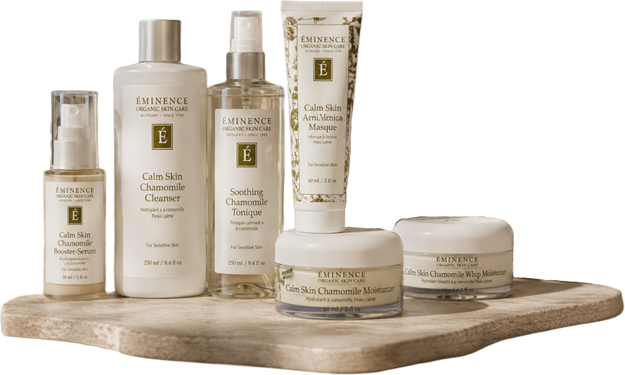 Collection of Eminence Organic Skin Care products, including Calm Skin Chamomile Cleaner, Booster-Serum, Tonique, Masque, Moisturizer, and Whip Moisturizer, arranged on a wooden surface.