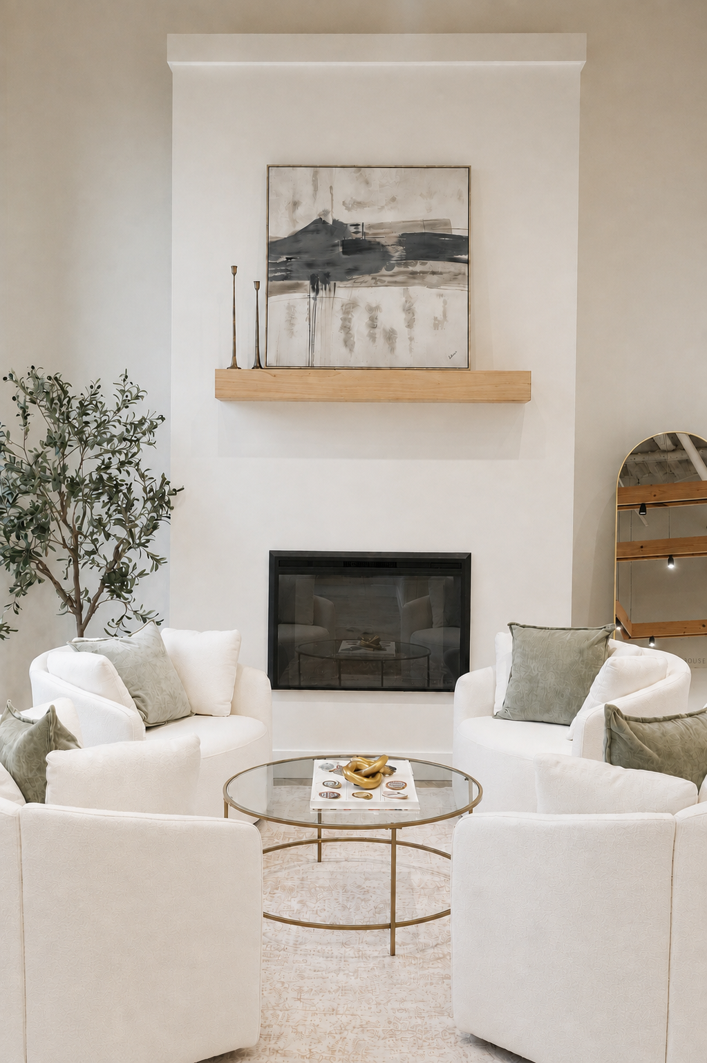 A cozy living room with a fireplace, four white armchairs with gray pillows arranged around a glass coffee table, a potted plant on the left, abstract artwork above the fireplace, and a wooden shelf with a mirror on the right.