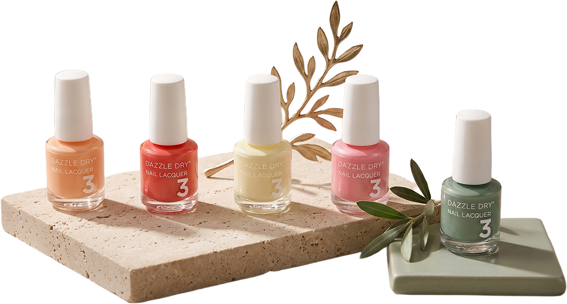 Five bottles of Dazzle Dry nail lacquer in peach, coral, yellow, pink, and green, arranged on a textured beige surface with decorative leaves around them.