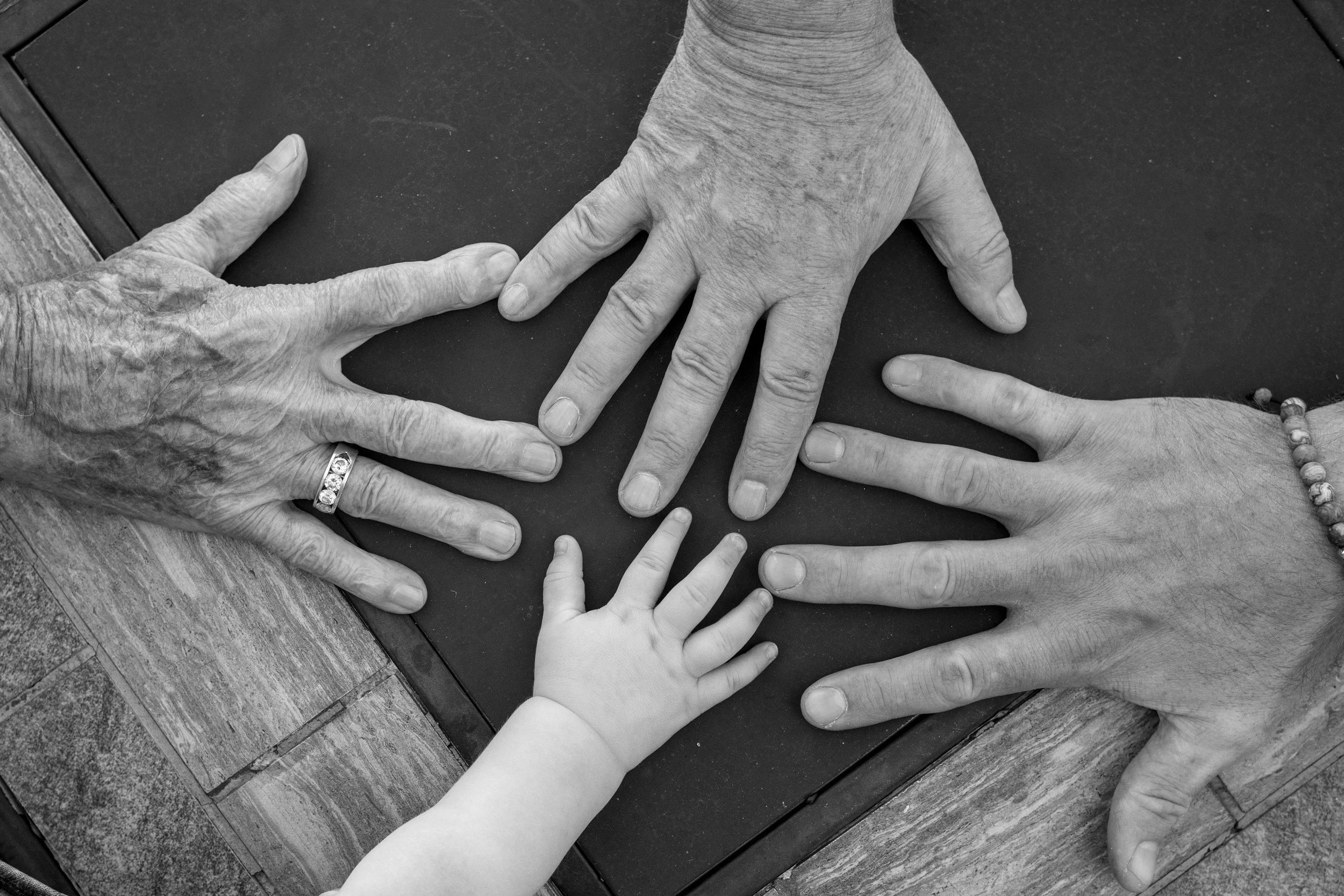 Multiple hands, including elderly and young children, are placed on a dark table, symbolizing connection or unity.