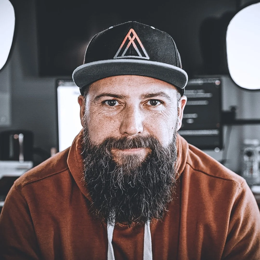 Close-up of Brian Crum, founder of MensMinistry.online — a man with a full beard wearing a black baseball cap with a geometric logo, brown hoodie, sitting in a tech office or studio environment.