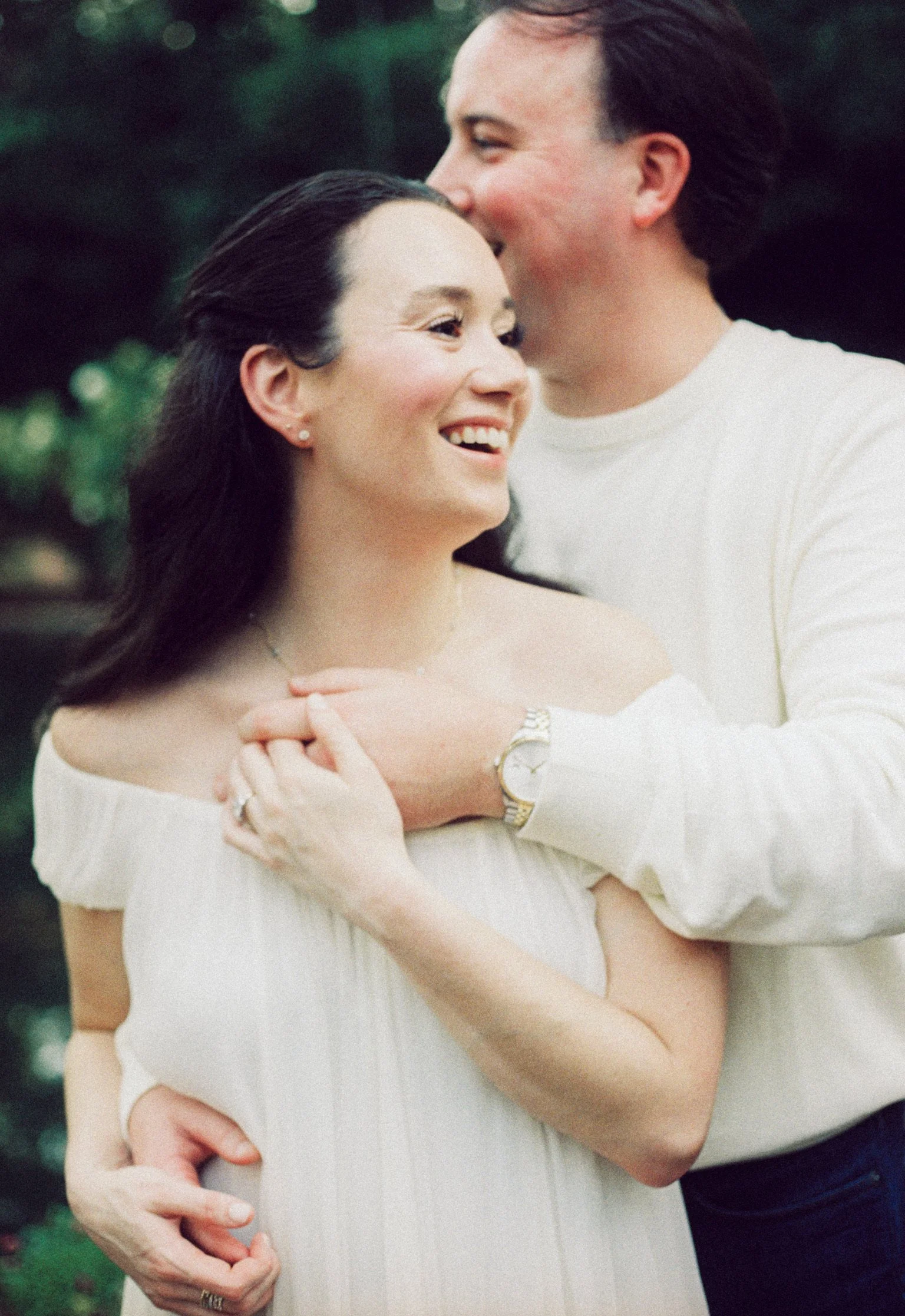 A couple, with the man whispering in the woman's ear, standing outside with greenery in the background. Both are smiling and appear happy.