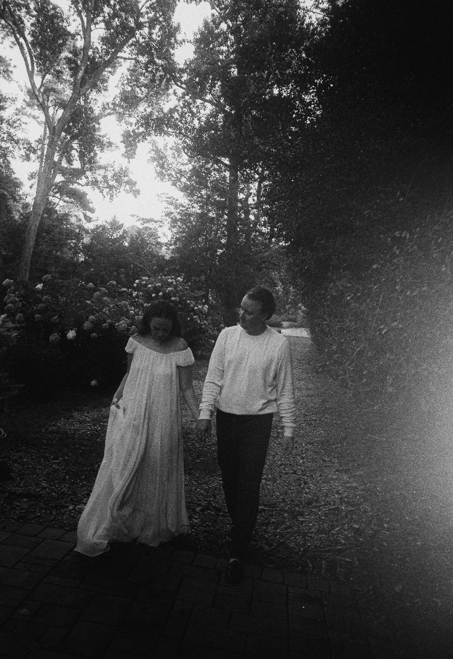 A black and white photo of a young man and woman walking outdoors. The woman wears a long, striped dress, and the man wears a long-sleeved shirt and dark pants. They are holding hands and looking at each other. The background has trees and bushes.